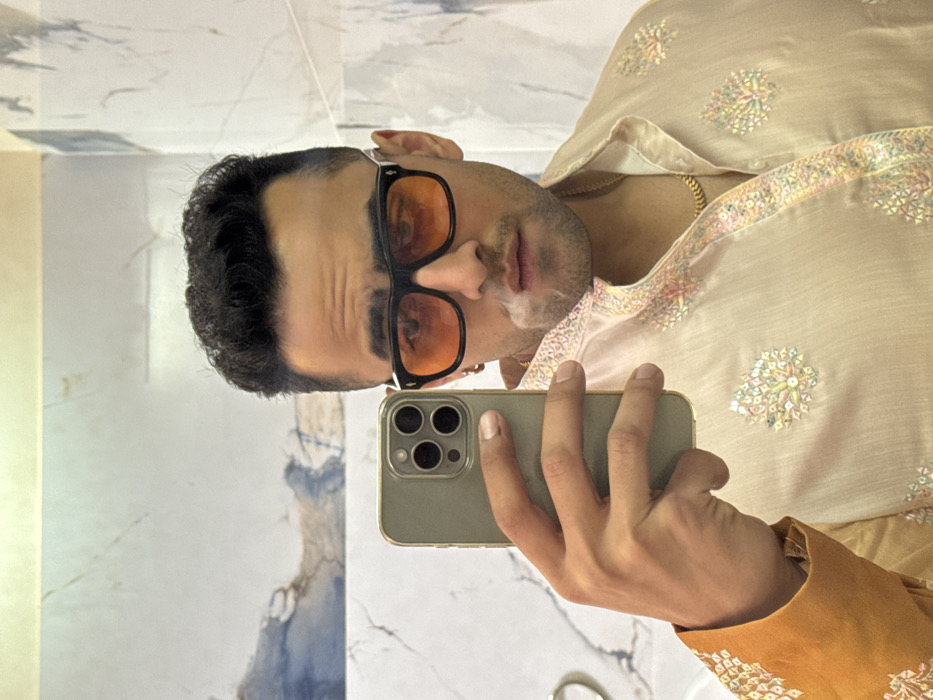 A male individual is captured in a mirror selfie within an indoor setting, likely a bathroom, indicated by white and grey marble-patterned wall tiles and a partially visible silver fixture on the left. The subject, who has dark hair and is wearing black-framed sunglasses with orange-tinted lenses, a gold chain, and a light-colored traditional Indian garment with intricate embroidery, is seen exhaling a plume of smoke from his mouth. He is holding a light-colored smartphone with a prominent triple camera array in his right hand to capture the image. A contrasting orange sleeve with embroidery is visible on the left side of the frame. The precise location within the city of Malasar, India, cannot be determined from the visual evidence.