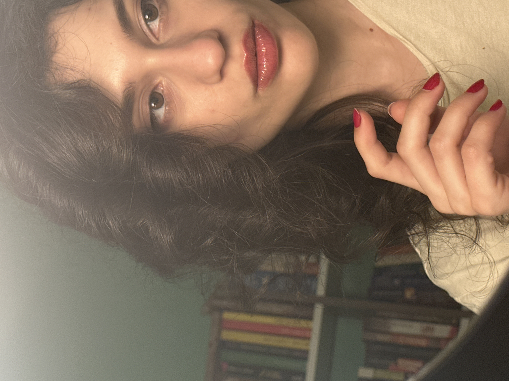 The image shows a close-up of a young woman's face, with her hair partially obscuring her left side. She has dark, curly hair and brown eyes. Her lips are painted with a glossy, reddish lipstick, and her fingernails are painted with a matching red polish. She is wearing a light-colored t-shirt. In the background, a bookshelf filled with books is visible, and the wall behind it is painted a muted green. The lighting suggests an indoor setting, possibly a home or a study. The overall impression is of a candid portrait, with the woman looking slightly away from the camera. There is no indication of the city of Sofia, Bulgaria in the image or its context.