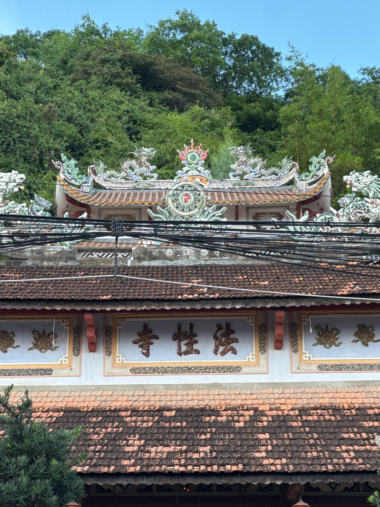 The image shows a tiered, ornate building roof adorned with colorful dragon sculptures and decorative tiles. Behind the roof, a dense green hillside is visible, suggesting a natural or park-like setting. The foreground features an older, tiled roof, partially obscured by a dark green tree on the left. Overhead, a tangle of black electrical wires spans across the frame, crossing in front of the ornate upper roof. Below the upper roof, a section of wall displays prominent East Asian characters painted in a deep red hue, enclosed within decorative rectangular panels. The overall scene depicts a structure with traditional architectural elements set against a lush green backdrop, with modern utility infrastructure crossing the view. This appears to be a temple or similar cultural site, possibly located near a natural elevation within Nha Trang, Vietnam, given the vegetation and architectural style.