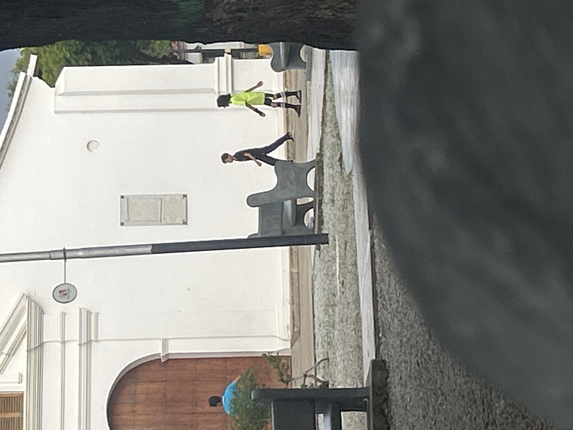 The image shows a street scene in Santa Ana, Venezuela, with a white building and two people walking. The building appears to be a church or other public institution with a large wooden door and architectural details. The ground in front of the building is covered with gravel. There are two benches visible, one in the foreground and another further back. A street lamp is also present. One person is wearing a blue shirt and walking towards the building. Another person, a child, is wearing a bright yellow shirt and dark shorts, walking away from the building. The scene is outdoors during the day.