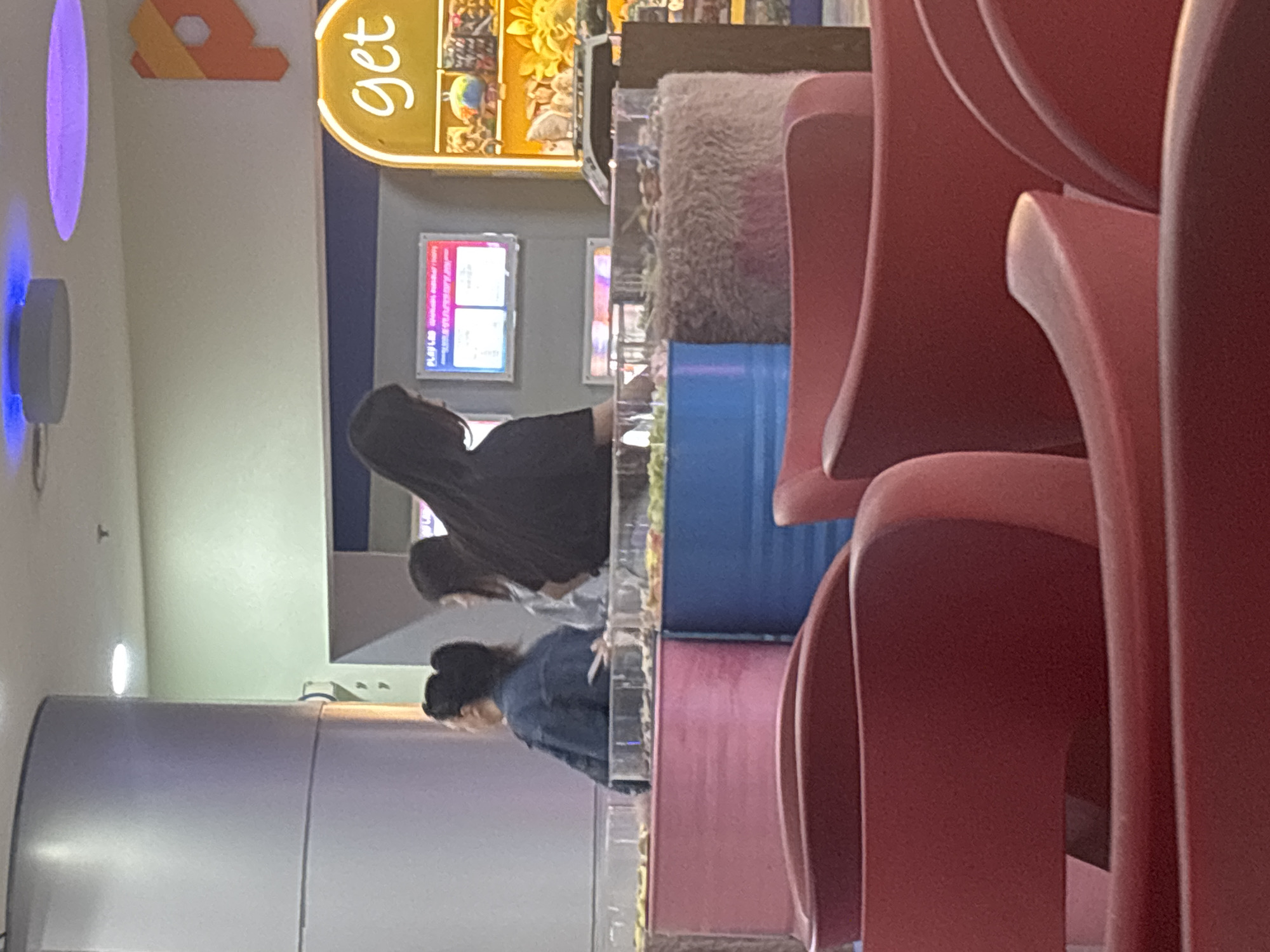 This image captures an indoor scene within a commercial or entertainment establishment, likely in Yerevan, Armenia. The foreground is dominated by a series of red, scoop-style plastic seats, suggesting a waiting area or audience seating.
In the mid-ground, three individuals are seen from behind, facing away from the viewer. The closest person wears a dark top, the second a lighter shirt, and the third, further to the left, is in a denim jacket with hair tied up. They appear to be at a counter or barrier, looking towards displays or activity stations.
The background features various elements. On the back wall, two brightly lit rectangular screens show colorful graphics and some text, with "PLAY LAB" partially visible on one. Above these screens and slightly to the right, a prominent yellow, oval-shaped sign reads "get" in white script. To the right of the screens, a display area holds various items, including a large yellow sunflower-like decoration and a globe, possibly prizes or merchandise. Below this display, colorful cylindrical objects, one blue and one pink, are stacked, possibly as part of the decor or a game setup. A furry, light-colored texture is also visible near these cylinders.
On the far left side of the scene, a large, light-colored cylindrical structure stands, and above it, a wall-mounted device emits a distinct blue glow. The space is illuminated by artificial indoor lighting, and no external weather conditions are visible. The overall impression is that of an active, public venue, possibly an arcade or a game redemption area, with the people potentially engaged with games or waiting. Emotions of the subjects are not discernible as they are seen from the back.