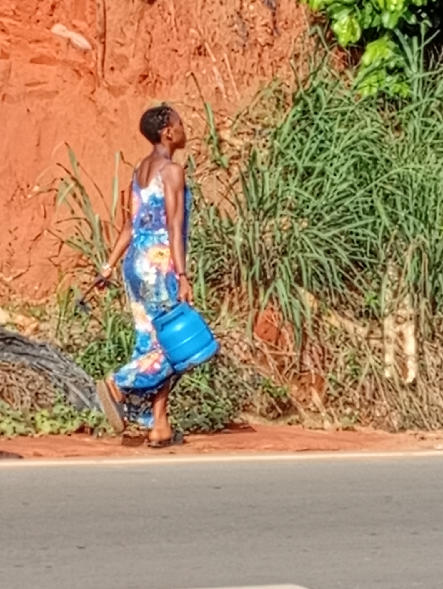 In Nkpor, Nigeria, a woman is walking along the road. She is wearing a floral dress and sandals. She is carrying a blue gas cylinder. The background consists of a red clay wall and overgrown green vegetation. The road is asphalt.