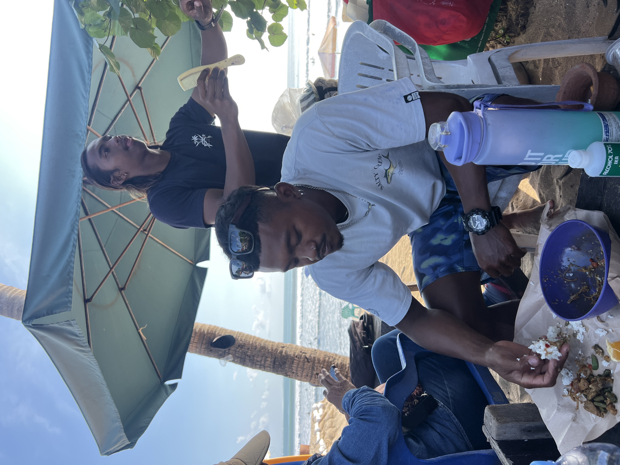 The image captures a casual outdoor scene on a beach, likely in Kuta, Indonesia, with the ocean and waves visible in the background. Two men are the primary subjects. One man, in the foreground, is seated and wearing a light-colored t-shirt with "SALTY CREW" printed on it, along with blue and white patterned shorts. He has dark skin, short dark hair, earrings, and a nose ring. He is actively eating from a brown paper spread with food, holding a piece of white rice in his hand, and a purple bowl is beside him. He wears a black wristwatch.
Another man stands behind him, partially obscured by a large, green beach umbrella. He has long dark hair, is wearing a black t-shirt with a graphic print, and is holding a yellow comb in his right hand. He appears to be interacting with the person in the foreground or another unseen individual.
The environment includes a sandy beach, a palm tree, a large green umbrella providing shade, and plastic chairs. Several objects are present, including a large purple water bottle with a blue and green gradient, a bottle of "ALCOHOL 70%," and a small mortar and pestle. Beach umbrellas and what appear to be surfboards are visible in the distance by the water, suggesting a typical Kuta beach setting. The overall atmosphere is relaxed and informal.