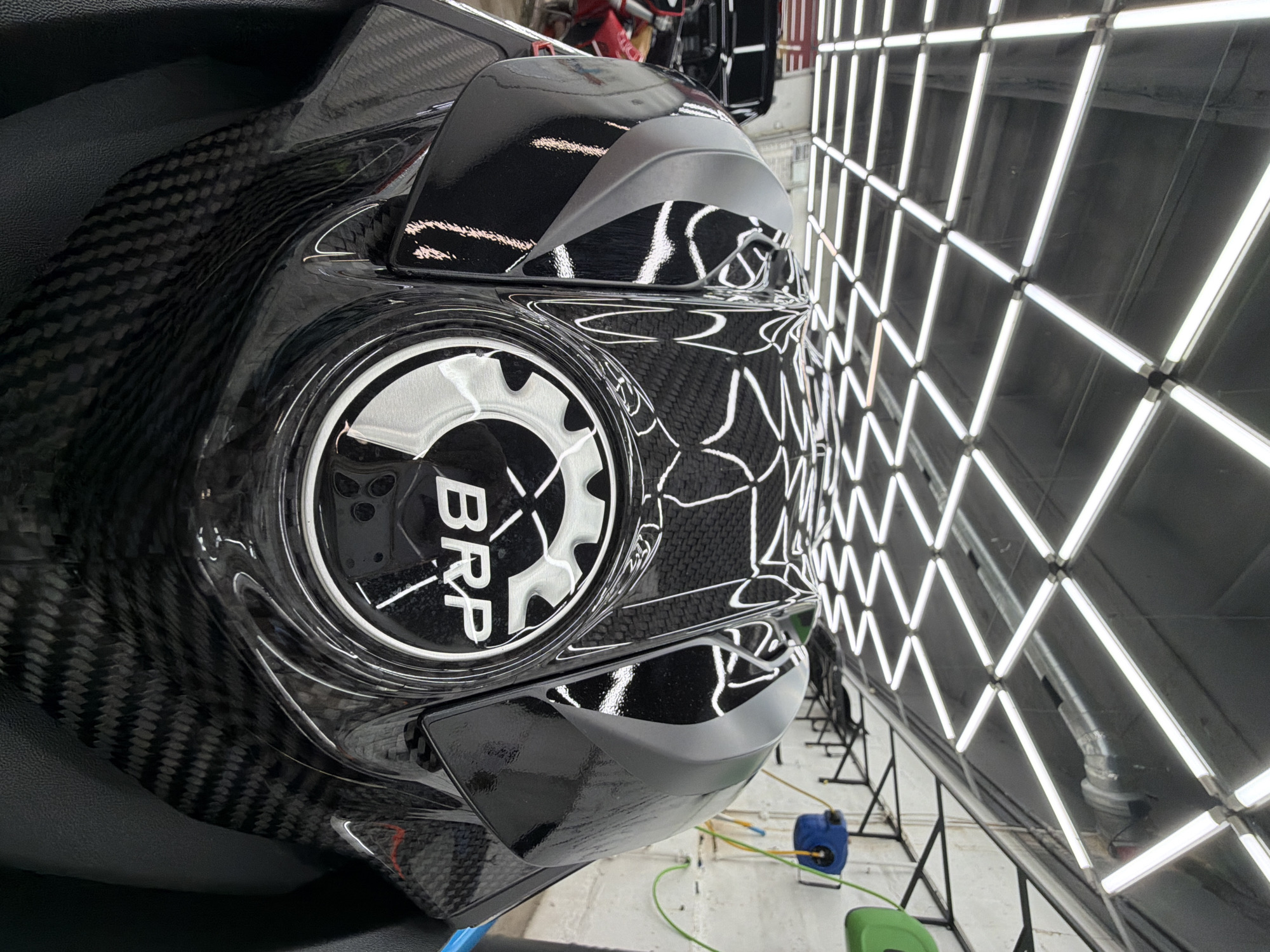 A close-up shot shows part of a carbon fiber vehicle, likely a snowmobile or ATV, bearing the "BRP" logo. The vehicle is positioned indoors, possibly in a detailing shop or showroom. The background features a wall lined with bright, white fluorescent lights arranged in a grid pattern. The environment includes cleaning supplies, like a blue canister and green container, positioned on the floor below the lighting structure. The location is unknown.