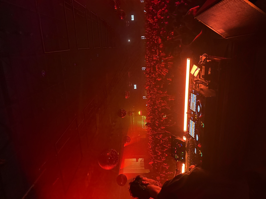 The scene depicts an interior view of a large, active music venue or nightclub located in Los Angeles, California, United States of America. The entire environment is saturated with intense red ambient lighting, augmented by a noticeable atmospheric haze. In the foreground, a lone individual, identified as a Disc Jockey (DJ) by their position and equipment, is visible from the rear perspective. This individual, possessing dark, curly hair, is positioned behind a professional DJ console comprising two media players and a central mixer unit, all illuminated by an orange LED light strip. To the right of the console, a dark, textured speaker cabinet is partially visible. The midground and background are occupied by a densely packed crowd of several hundred individuals. Their forms are largely silhouetted and indistinct under the dominant red light, with numerous arms raised in various gestures. Multiple large, reflective disco balls are suspended from the dark, high ceiling, reflecting the red light. Two illuminated signs, each displaying the word "BAR" in white capital letters, are visible above the crowd at different points within the venue, indicating service areas.