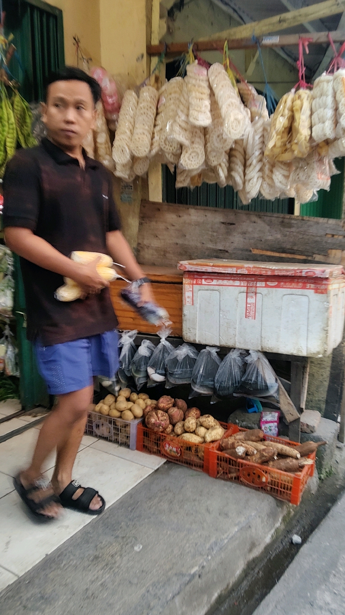 A man stands in front of a market stall in Mekarmukti, Indonesia, holding three ears of corn in his left arm and a plastic bag in his right hand. He is wearing a black t-shirt and blue shorts, with black sandals on his feet. The stall is filled with various produce, including potatoes, sweet potatoes, and cassava, displayed in crates. Bags of other goods hang from the ceiling, and a cooler is visible behind the man. The scene appears to be outdoors, on a paved street, with a building in the background.