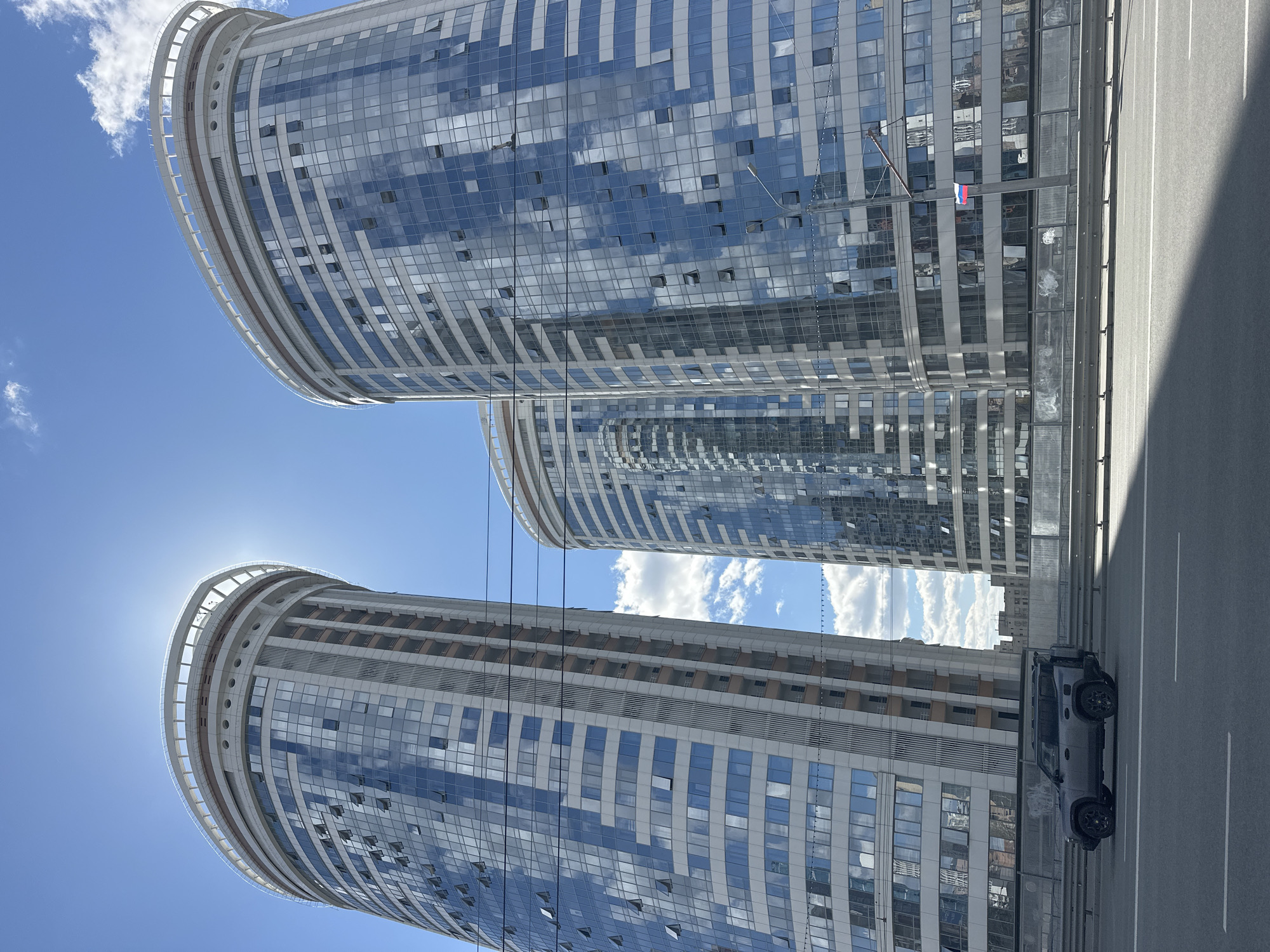 The image shows three modern, high-rise buildings in Novosibirsk, Russia. The buildings have curved, glass-covered facades that reflect the blue sky and white clouds. A road with a grey SUV is visible in the foreground. A Russian flag is visible hanging from one of the buildings. The sun is bright and partially obscured by the top of one of the buildings.