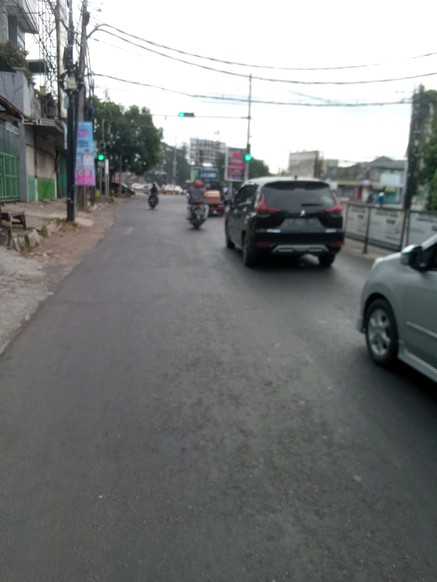 The image depicts a street scene in Jember, Indonesia. A black SUV is driving on the road, followed by a light-colored car. In the background, there are traffic lights, buildings, and trees. The sky is overcast. There are several motorcycles on the road, some of which appear to be moving. The road is asphalted and appears to be in good condition. The overall impression is of a typical urban street scene in Indonesia.