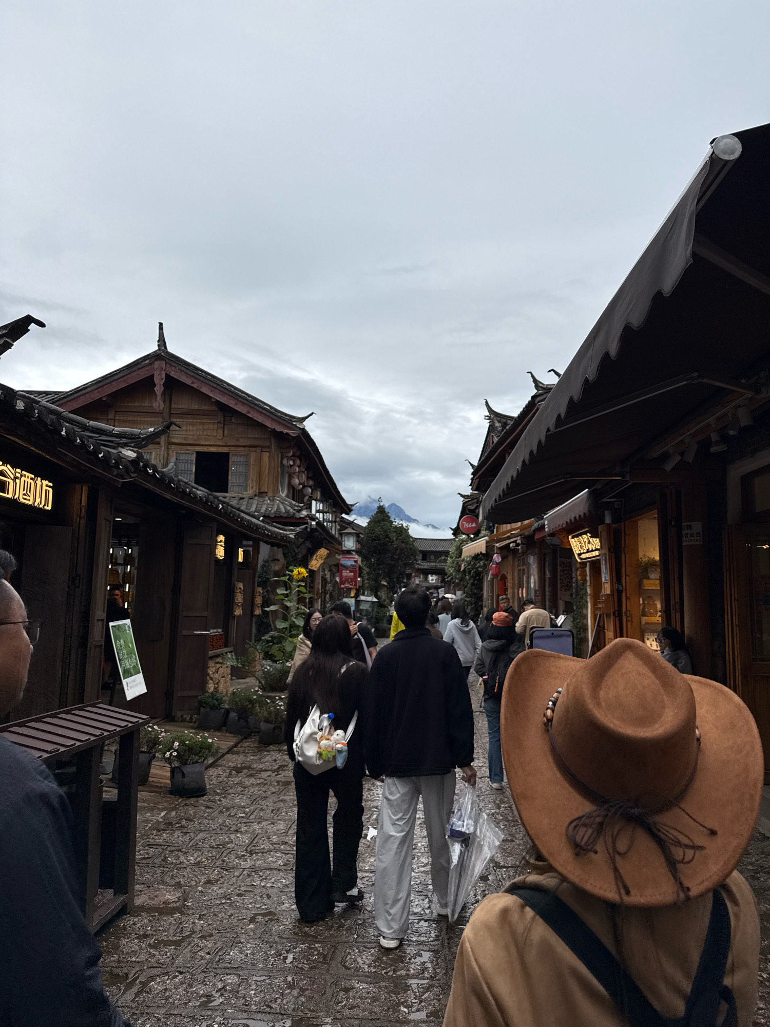 An outdoor, eye-level, medium shot captures a view from behind a person wearing a brown cowboy hat as they walk through a cobblestone alleyway in Yulong Nakhi Autonomous County, China. The alley is lined with traditional wooden buildings with dark tiled roofs. Several people are walking in the same direction, some carrying bags and an umbrella. The sky is overcast and the cobblestones appear wet, suggesting recent rain. The architecture and cobblestone street indicate a historic or culturally preserved area within Yulong Nakhi Autonomous County.