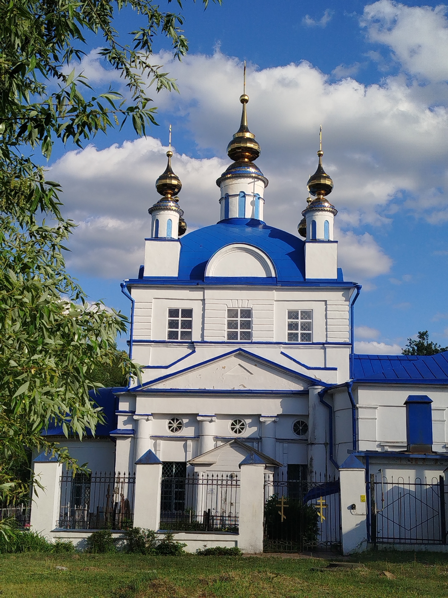 The image depicts a white and blue Orthodox church in Мамонтово, Russia. The church features three golden domes with spires, a blue roof, and white walls with blue trim. The building is surrounded by a black metal fence and some vegetation. A large leafy tree partially obscures the left side of the church. The sky is blue with scattered white clouds. The ground is covered with green grass.