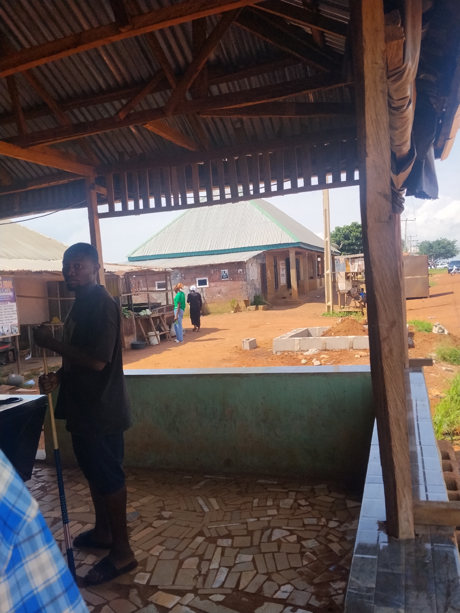 In the city of Durumi, Nigeria, an outdoor scene unfolds beneath a corrugated metal roof supported by wooden beams. A dark-skinned man, wearing a dark t-shirt and shorts, stands with a long pole in his hands, looking towards the camera. He wears black sandals. To his right, a person in a blue and white checkered shirt is partially visible. The ground beneath them is paved with irregularly shaped tiles. In the background, a low, green wall separates the foreground from a dusty, orange-colored ground. Several buildings with corrugated metal roofs are visible. One building, closer to the background, has white trim around its roof and a pale green facade. To the left of this building, a woman in a green shirt and blue jeans, and another person in a dark garment, walk away from the viewer. Further in the distance, trees and power lines are discernible under a bright, hazy sky.
