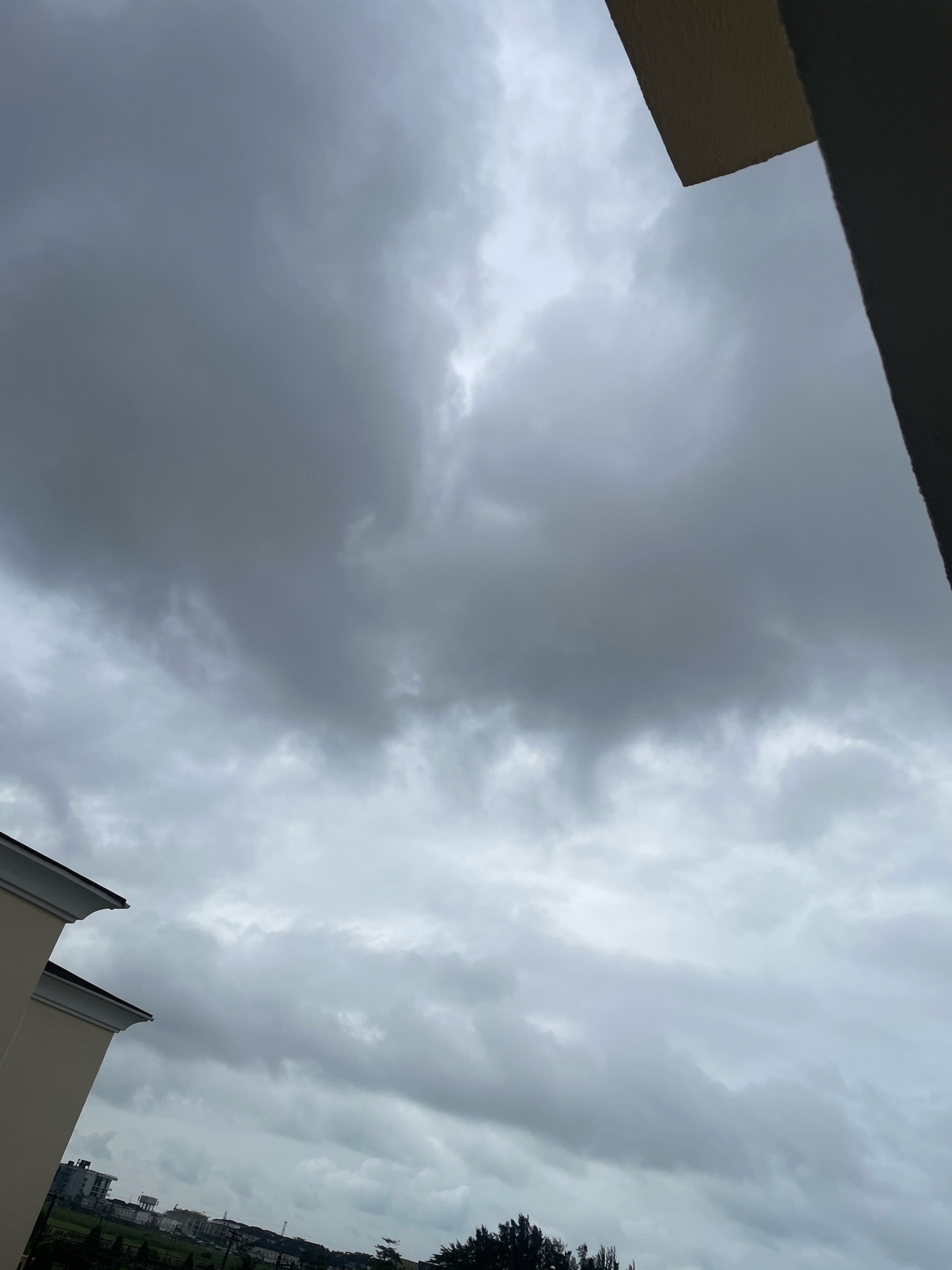 The image shows a cloudy sky over Lekki Peninsula II, Nigeria. The clouds are dark and heavy, suggesting an impending storm. Two buildings, possibly residential, are visible in the lower left and upper right corners of the image, with architectural features such as rooflines and walls. In the background, a low-rise cityscape with some trees and what appears to be a water tower can be seen, indicating an urban or suburban environment. The overall scene is dominated by the overcast sky and the muted colors of the buildings and landscape.
