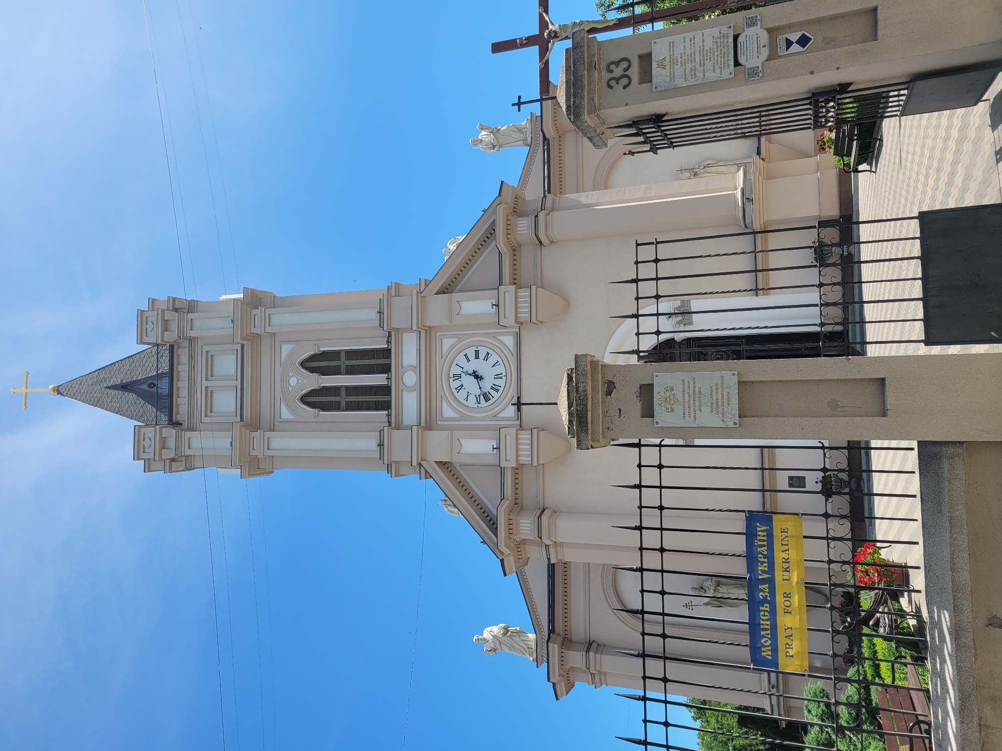 The image shows the facade of a church with a tall bell tower topped with a golden cross. The church is painted a pale pink color and features classical architectural elements, including columns, arches, and statues of religious figures. A large clock face is visible on the bell tower. In front of the church, there is a black metal fence. A banner with the Ukrainian flag colors and the text "MOLUS' ZA UKRAÏNU" (Pray for Ukraine) and "PRAY FOR UKRAINE" is hung on the fence. To the right of the entrance, a pillar with the number "33" and a sign with Ukrainian text is visible. A crucifix with a statue of Jesus Christ is also present near the entrance. The sky is clear blue, and the scene appears to be bathed in sunlight.