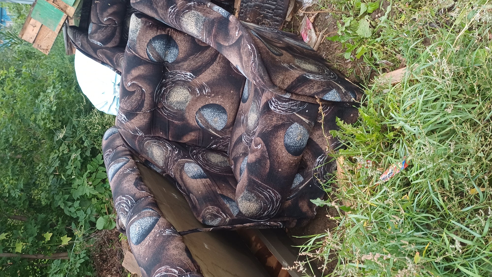 An old, upholstered armchair with a brown and blue circular pattern is situated outdoors amongst overgrown green vegetation and debris. The armchair appears weathered and possibly discarded. In the background, there is dense foliage, trees, and what appears to be stacked lumber. A white object, possibly a blanket or tarp, is partially visible behind the armchair. A small, colorful piece of debris, possibly plastic or paper, is visible in the grass in the foreground. The scene suggests an outdoor, unkempt area, possibly a backyard or a waste disposal site in Izhevsk, Russia.