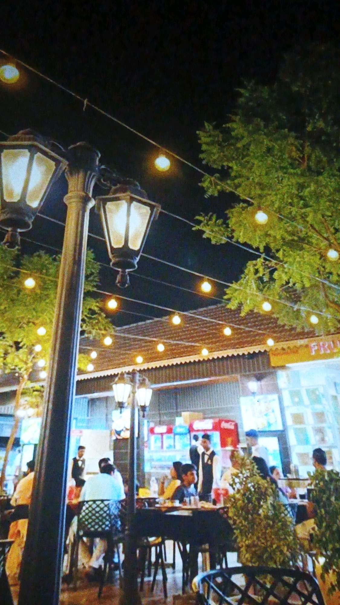 This is a photograph of an outdoor dining area at night. The scene is illuminated by decorative string lights and a large lamppost in the foreground. Trees with green foliage are visible, as are tables and chairs set up for customers. Several people are seated at the tables, and at least two individuals, possibly servers, are standing. In the background, there appears to be a building with signage. One sign partially visible reads "FRU." The overall atmosphere suggests a restaurant or food establishment. The presence of "FRU" on the signage and the general ambiance could be indicative of a location like "Fruit" or a similar establishment in Indore, India, known for its open-air dining experiences.