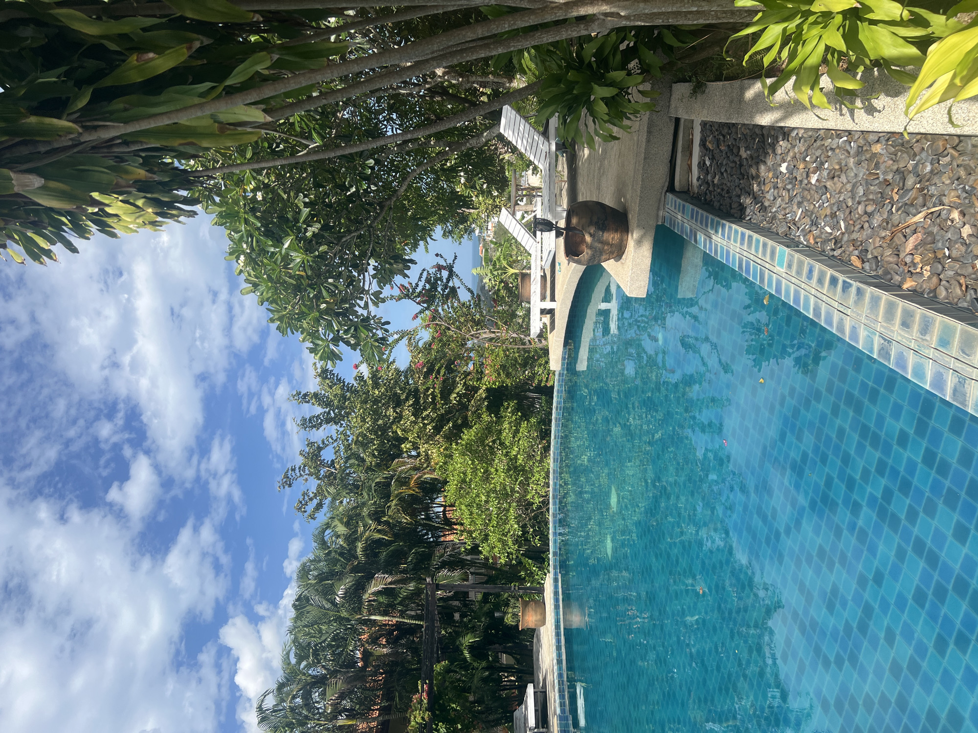 The image depicts a poolside scene in the city of Ko Samui District, Thailand. In the foreground, a swimming pool with blue tiled lining and clear turquoise water occupies the left side of the frame. The pool's surface reflects the sky and surrounding foliage. Along the edge of the pool, a walkway paved with stone leads to two white lounge chairs. A large, dark, ceramic pot, possibly a planter or decorative item, sits on the walkway near the lounge chairs. The background is filled with lush tropical vegetation, including palm trees and other green plants, with a glimpse of the ocean and distant buildings visible through the trees. The sky is blue with scattered white clouds, suggesting a clear, sunny day. There are no people visible in the image.