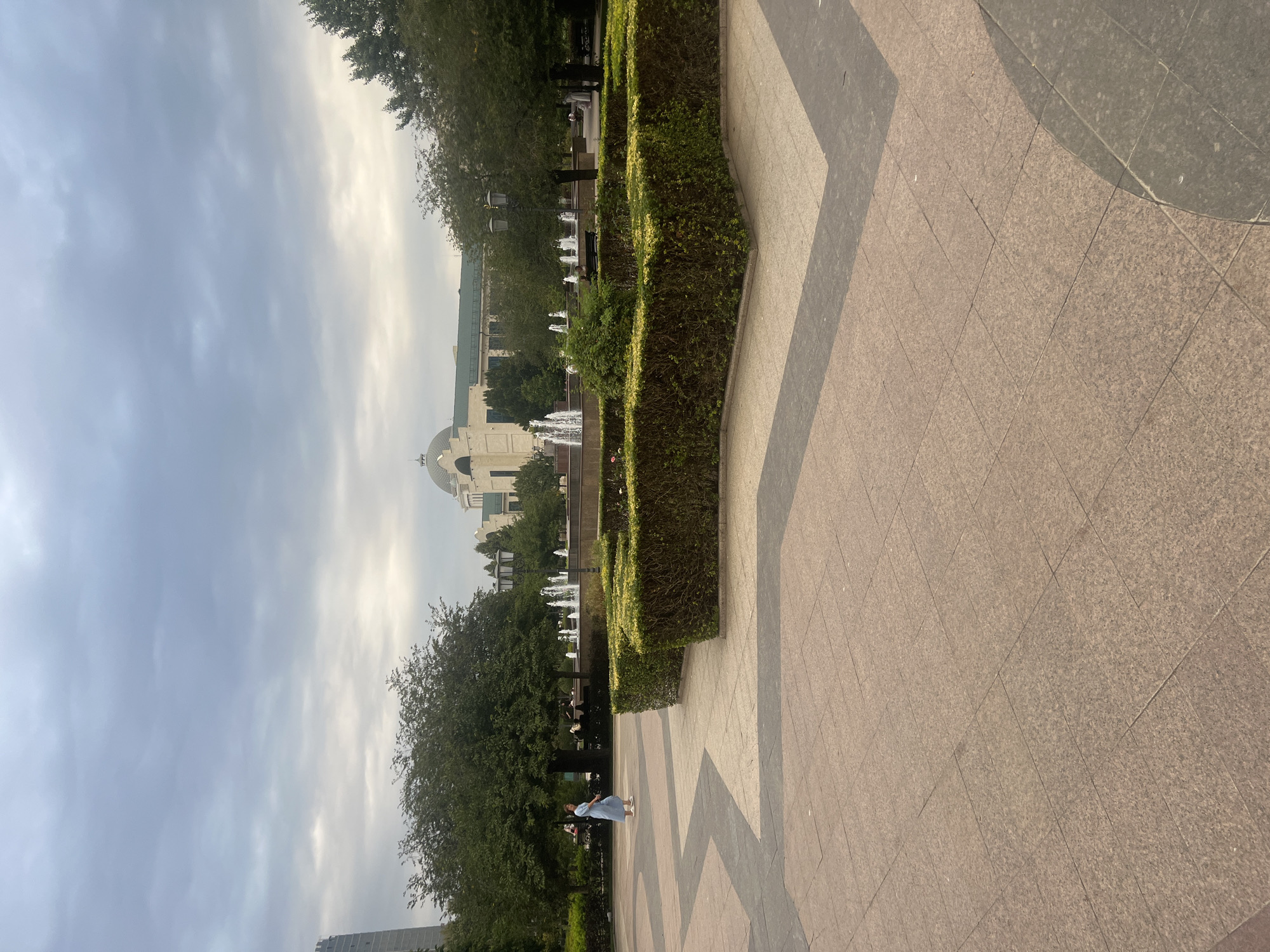 A woman in a light blue dress walks across a paved plaza in Grozny, Russia. The plaza features a geometric pattern of light and dark paving stones. In the background, a neatly trimmed hedge forms a low garden bed, behind which several fountains spray water into the air. Beyond the fountains, trees line a park area with benches, and further in the distance, a large building with a domed roof and green trim is visible. The sky is overcast.