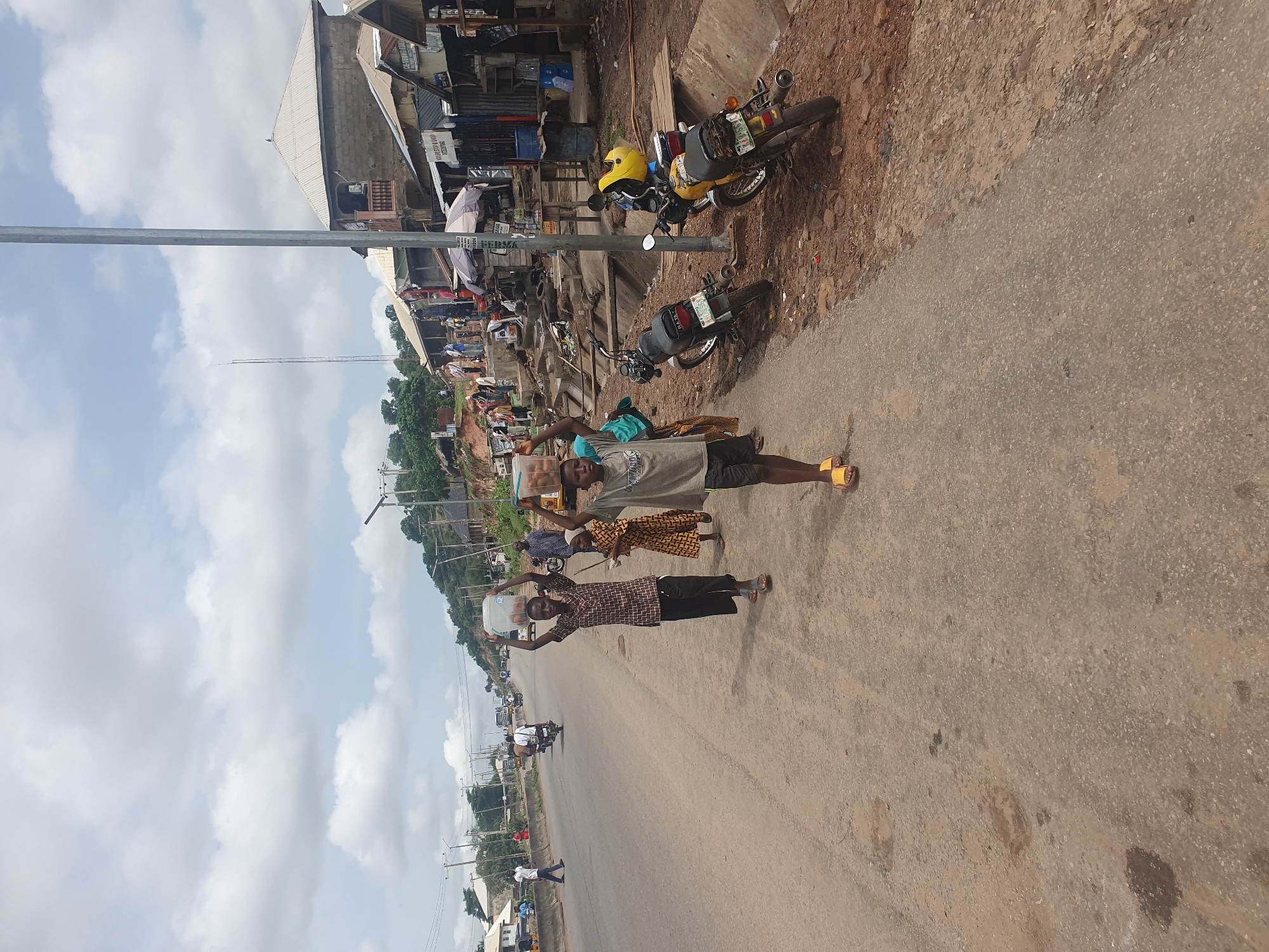 The image shows a street scene in Lokoja, Nigeria. Three people are carrying items on their heads while walking along the side of a road. The first person is carrying a plastic container of water. The second is carrying a package. The third is carrying a carton of eggs. There are motorcycles parked on the side of the road along with building and general wares. The sky is partly cloudy. Motor vehicles are visible in the distance along the road.