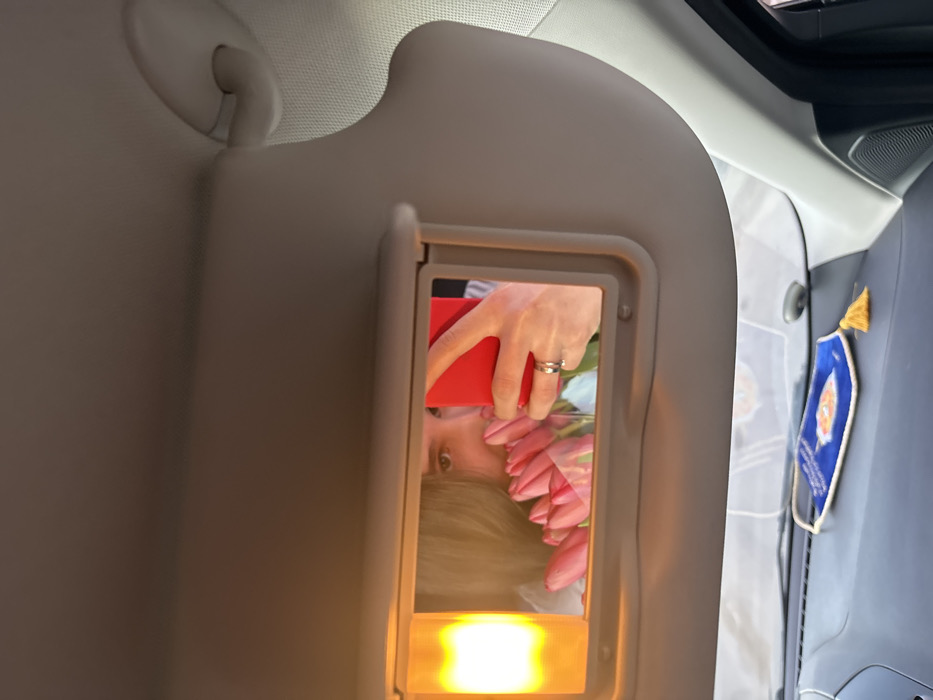 The image displays the interior of a vehicle, observed from a forward-looking perspective, presumed to be in the city of Slavyansk-na-Kubani, Russia. The primary subject is the driver's side sun visor, lowered to reveal an integrated vanity mirror with an illuminated orange-yellow light source on its left. Reflected in this mirror is a person, partially visible, holding a bouquet of pink tulips wrapped in clear plastic near their face. The person's right hand, adorned with a silver-colored ring on the ring finger, holds a vibrant red smartphone, angled as if capturing a selfie. The person's long, light brown hair is also visible in the reflection. Below the sun visor, a dark-toned dashboard is present. Resting on the dashboard is a blue fabric ornament, possibly a car charm, featuring an orange embroidered design and a yellow tassel. A small, dark, circular object is also discernible on the dashboard surface. The vehicle's ceiling and an overhead grab handle, both in a light beige or grey color, constitute the upper portion of the frame.