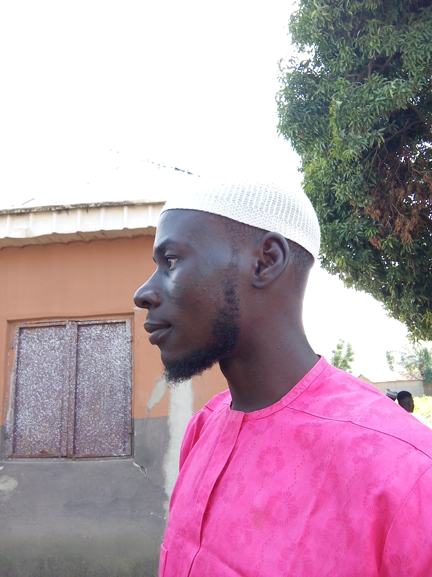 An outdoor, medium shot shows a Black man, from the chest up, in profile facing left. The man wears a white, crocheted prayer cap and a bright pink, long-sleeved garment with a floral pattern. His skin is dark, and he has a short beard. He stands before a light brown building with a white roofline and a closed, metallic window. To the right of the man, a large green tree dominates the background. The sky is overcast and bright.