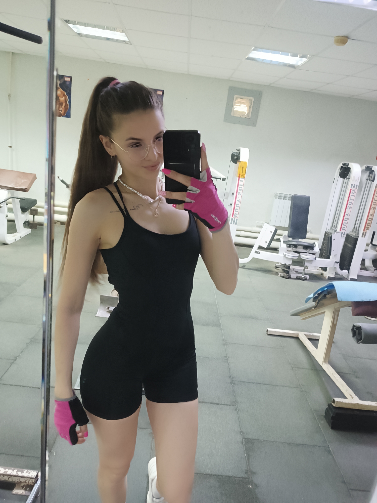 A woman is taking a selfie in a gym. She is wearing a black romper, pink workout gloves, and glasses. She has long brown hair in a ponytail. The gym has various weight machines and benches. There is a mirror on the left side of the image, and fluorescent lights are visible on the ceiling. The location is Unknown, Unknown.