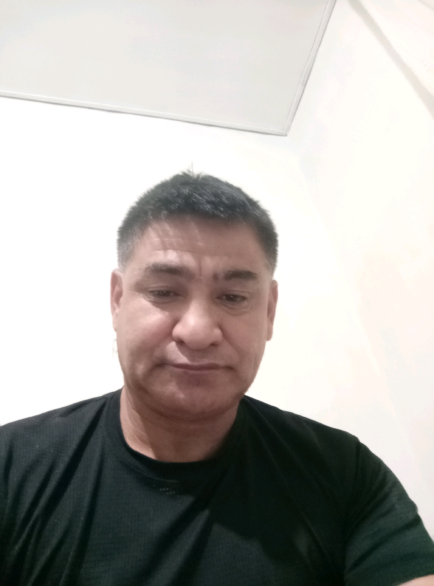 A head-and-shoulders, medium-shot photograph shows a man of Asian descent facing the camera. He is wearing a black t-shirt. The background is a plain, light-colored wall, with a white ceiling visible at the top of the frame. The lighting is diffuse and appears to be artificial. The image was taken in Hojakarzan, Uzbekistan.