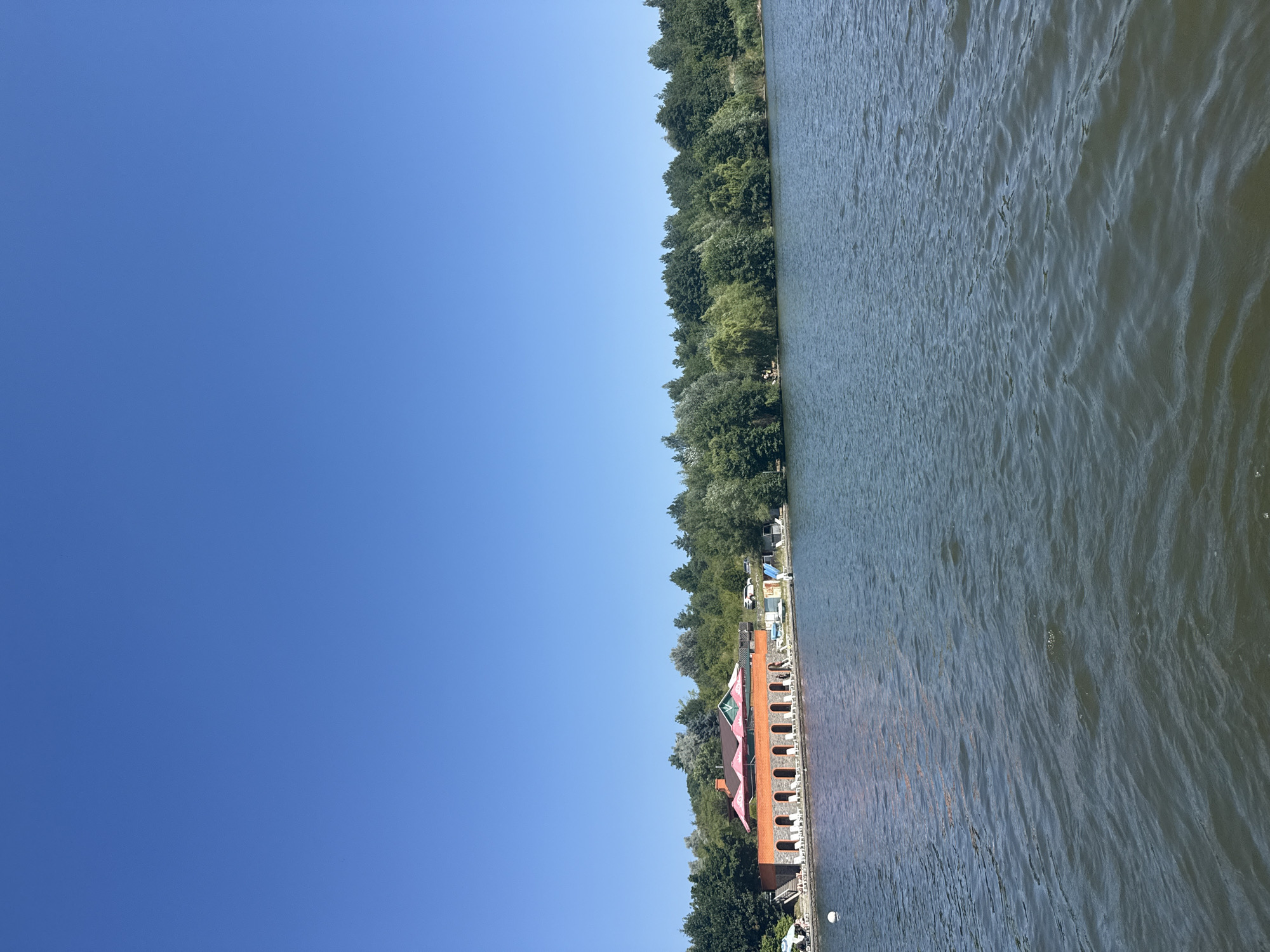 The image depicts a body of water with a resort-like establishment on the shore in Tiutky, Ukraine. The water occupies the lower half of the frame, showing gentle ripples and a dark, murky color. The shoreline features a long, orange-brick building with an arched facade, lined with white lounge chairs. Behind this building, a green-roofed structure is visible, and a line of lush green trees forms the backdrop. The sky above is a clear, deep blue. A few small boats are moored near the shore, and what appears to be a water slide is also present. The overall impression is a tranquil summer day at a lakeside resort.