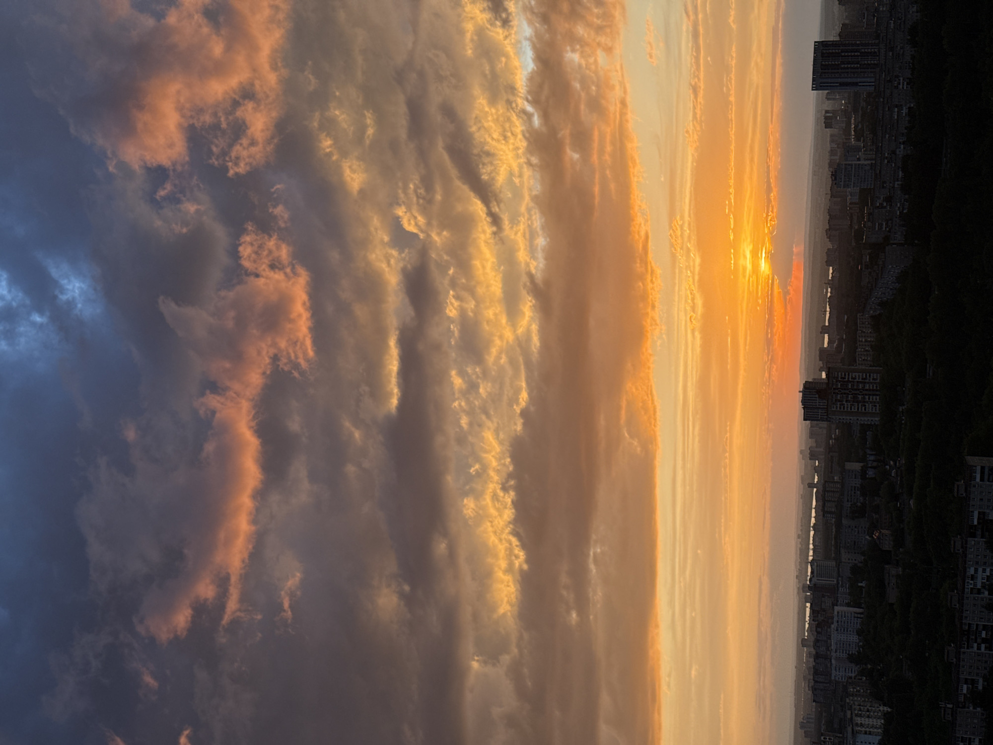 The image shows a sunset over the city of Perm, Russia. The sky is filled with clouds, ranging in color from dark blue and gray to orange and yellow. The sun is setting on the horizon, casting a warm glow over the city. In the foreground are buildings, likely residential and commercial, rendered in silhouette against the bright sky. The image is taken from an elevated vantage point.