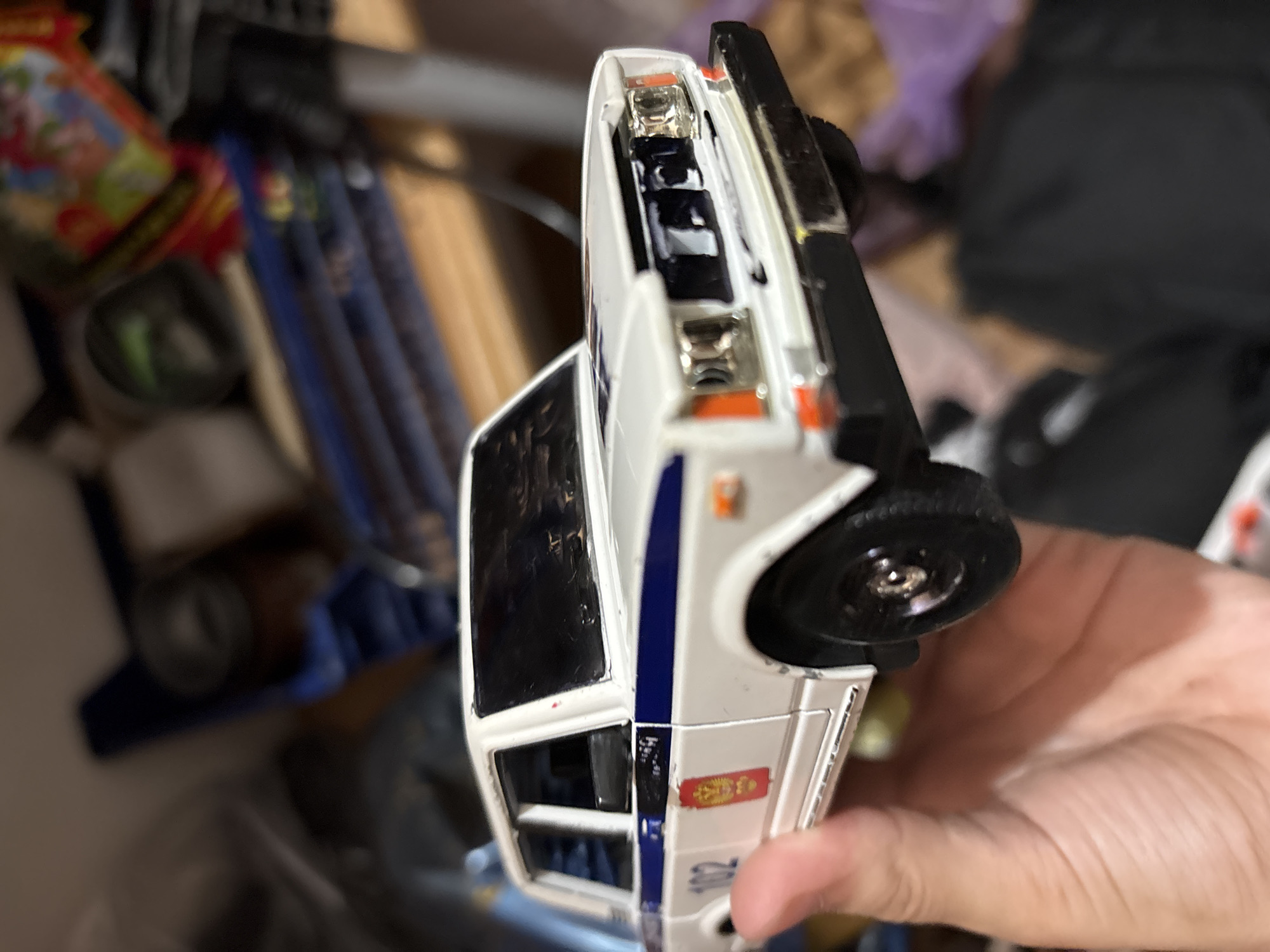 A white toy police car is being held up by a person's hand in front of a blurred background. The car is white with a blue stripe along the side and the number "102" is visible. There is a red insignia on the door. The background appears to be indoors with shelves and various objects that are out of focus. There is no discernible information about the location in the city of Grozny, Russia, within the image.