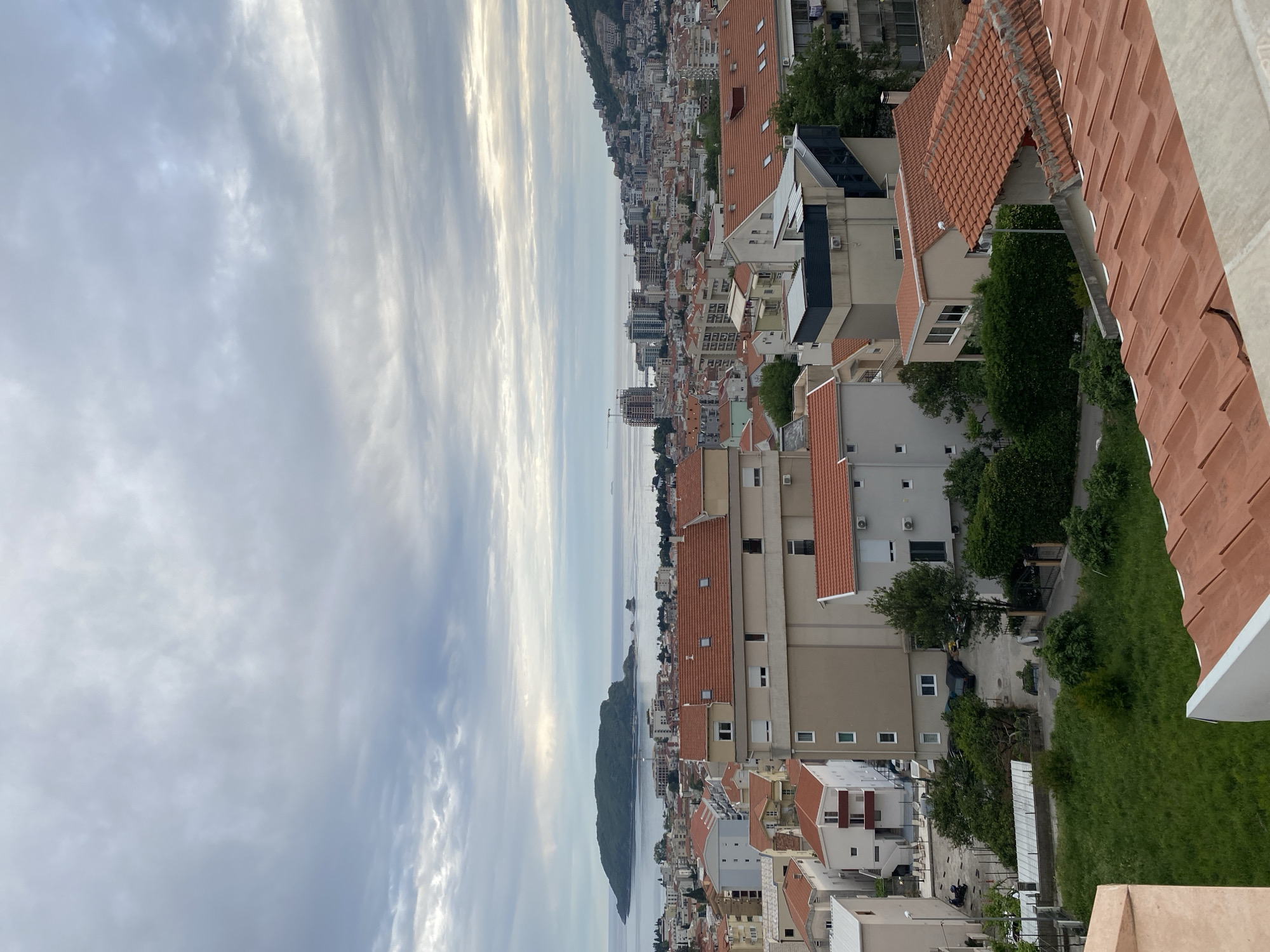 The image is an aerial view of the city of Budva, Montenegro, overlooking the Adriatic Sea. The foreground consists of a close-up of terracotta rooftops and some greenery. A dense concentration of buildings with similar terracotta roofs occupies the mid-ground, interspersed with trees. In the distance is the coastline stretching into the horizon, and a small island sits in the water. The sky is cloudy with patterns suggesting diffused sunlight. The scene appears to be static and unpopulated from this vantage point.