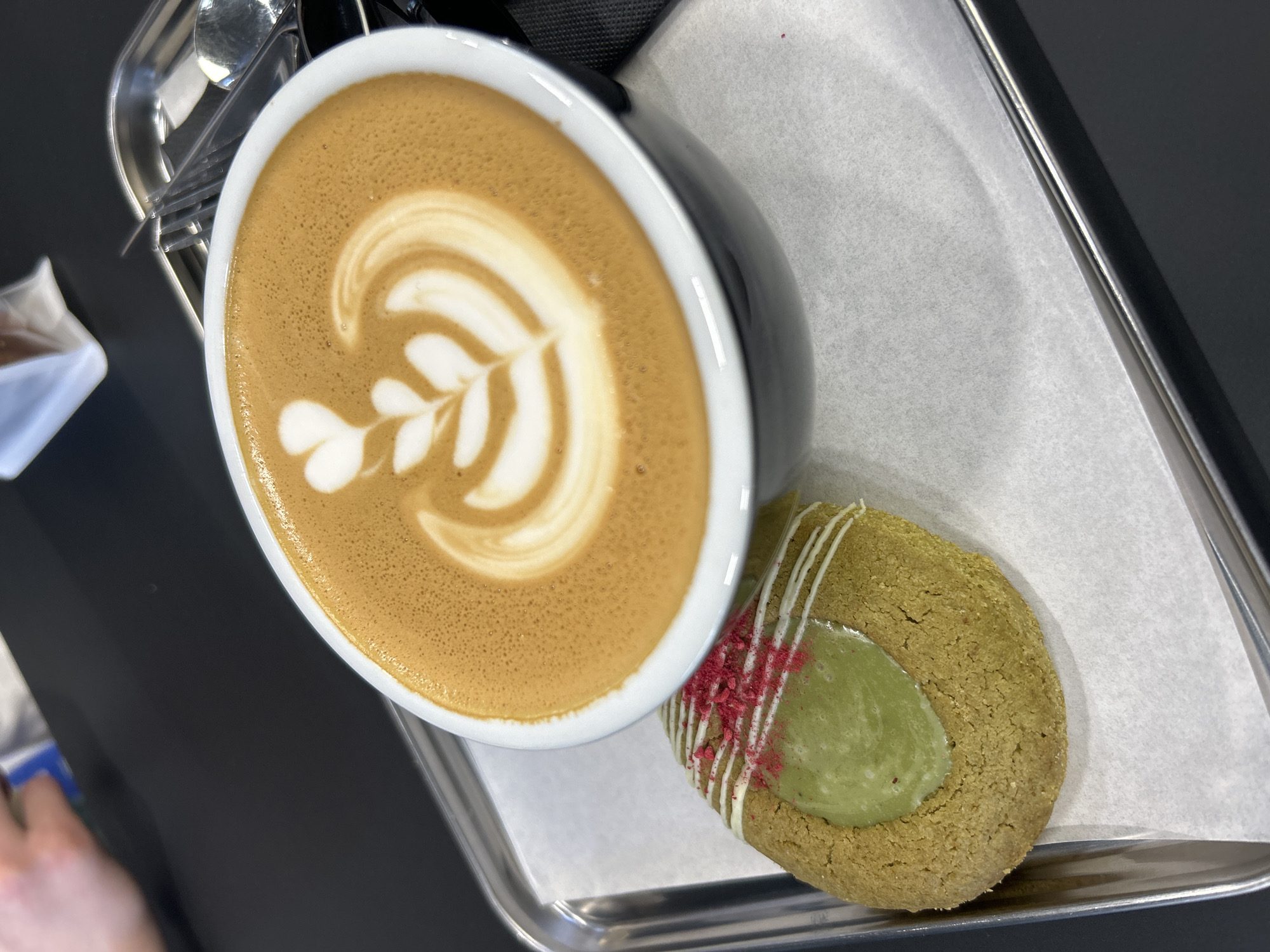 This overhead, close-up shot captures a coffee beverage and a decorative cookie presented on a metal tray. The scene suggests an indoor setting, likely a cafe or similar establishment, with the items resting on a dark, uniform surface that appears to be a table or countertop.
The main subject on the left is a dark-colored ceramic cup, possibly black or dark grey, with a white interior rim. It contains a latte or cappuccino, distinguished by elaborate white latte art featuring a stylized, multi-leafed design on the tan-brown surface of the coffee. The frothy texture of the drink is clearly visible.
To the right of the cup is a round, flat cookie. The cookie's base has a light, muted greenish-brown hue, possibly indicating a matcha flavor. Its center is filled with a smooth, lighter green frosting or glaze. The cookie is further adorned with thin, irregular lines of white icing drizzled across one side, which are then sprinkled with small, bright reddish-pink crumbs, likely freeze-dried berries.
The coffee and cookie are placed on a rectangular metal tray, which is lined with white, slightly textured parchment paper. A small portion of a metallic utensil is visible in the upper left background, and a blurred white object, possibly a sugar packet or napkin, is partially seen in the lower left. The lighting is bright and even, typical of an indoor daytime environment. There are no people visible, nor is there any discernible text within the image. The visual cues do not specifically indicate the location as Izhevsk, Russia, beyond the provided context, but the setup is consistent with a cafe experience.