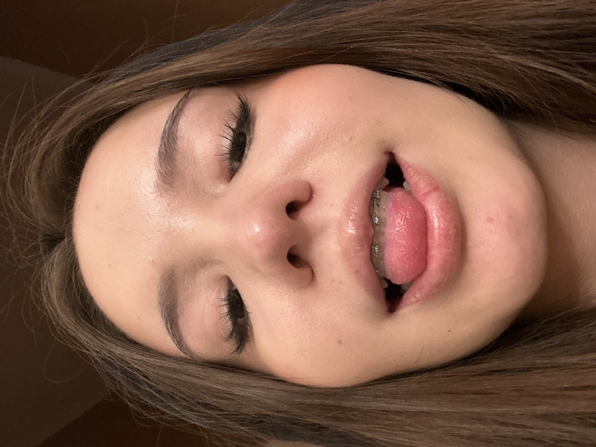 A close-up, low-angle portrait captures a young individual, likely female, with medium-brown hair framing the face. The subject's head is tilted slightly back, with eyes closed, revealing long, dark eyelashes. The nose is prominent, and the mouth is open, with the tongue partially extended. Braces are visible on the upper teeth. The lips appear glossy, and the skin exhibits a subtle sheen, with minor imperfections visible. The background is indistinct and dark brown, suggesting an indoor setting. This scene is located in the city of Naberezhnye Chelny, Russia.
