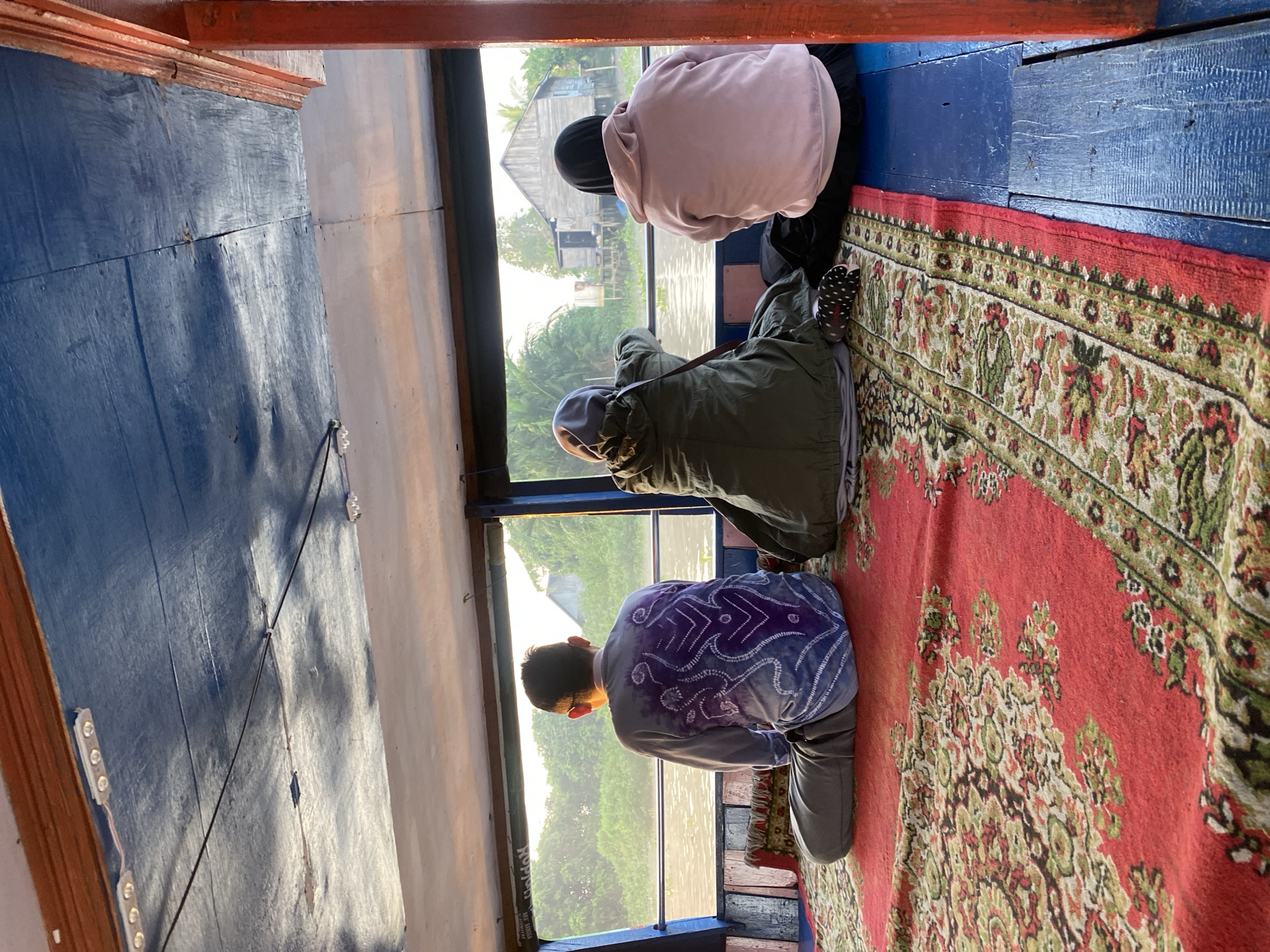 Three individuals are seated on a boat's floor, looking out a large window at a riparian landscape. The person on the left is a male wearing a grey long-sleeved shirt with a purple batik pattern and grey pants, sitting cross-legged. In the center, a person in a green hooded jacket and a light purple headscarf is seated. To the right, a person wearing a pink hooded sweatshirt and a black headscarf is also seated. The boat's interior features a blue ceiling with lights and wooden beams. A red oriental rug with a floral pattern covers the floor where the individuals are seated, transitioning to a blue painted wooden floor to the right. Outside the window, a river is visible with lush green vegetation along its banks, and a wooden house on stilts can be seen. The lighting suggests it is either early morning or late afternoon. This scene is consistent with the environment found in villages along rivers in Sungai Tandipah, Indonesia, where boat travel is common and houses are often built on stilts.