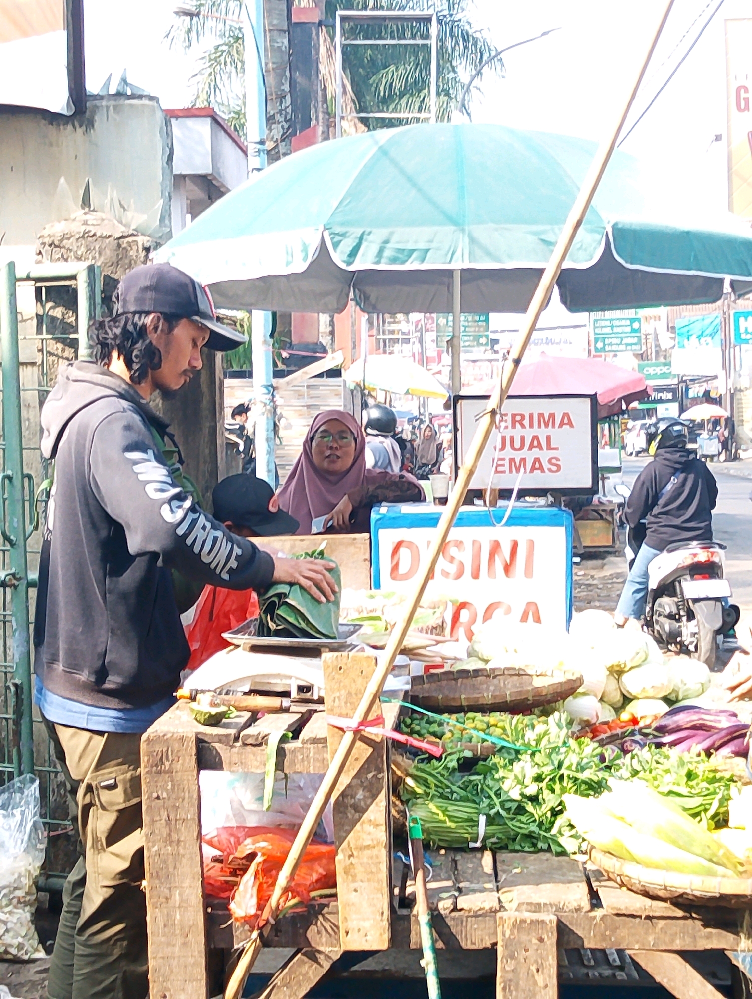 This image depicts a street market scene in Lembang, Indonesia, under a bright, sunny sky. In the foreground, a man wearing a black baseball cap and a black hooded sweatshirt with white lettering on the sleeve is standing at a wooden stall. He has long, dark, curly hair and is wearing olive-colored cargo pants. He is in the process of placing a bundle of items wrapped in green leaves onto a white digital scale. The stall is filled with various fresh produce, including bundles of leafy green vegetables, eggplants, and corn. Behind him, a woman wearing a maroon hijab and glasses is seated at the stall, looking towards the viewer.
A large, teal-colored umbrella provides shade over the stall. In the background, other market stalls and shoppers are visible, as well as motorcycles and vehicles on a road, indicating a bustling urban environment. Two signs are prominently displayed on the stall. One sign, in red lettering, reads "ERIMA JUAL EMAS" which translates to "Welcome Sell Gold". Another sign, in white with blue trim, says "DISINI" which means "Here". The overall impression is that of an active marketplace where goods, including vegetables and potentially gold jewelry, are being sold.