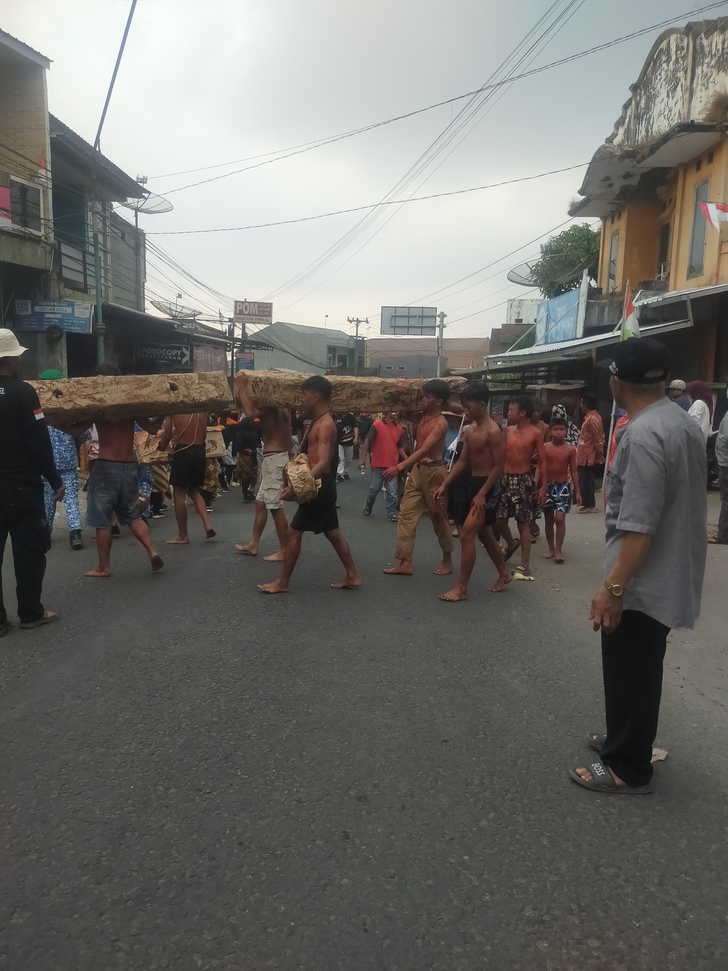 In Tuwel, Indonesia, a group of shirtless young men are carrying large, rectangular wooden beams on their shoulders as they walk down a paved street lined with buildings. The sky is overcast, and there are overhead power lines and satellite dishes visible. Some of the men are barefoot, and others are wearing shorts or light-colored pants. A man in a grey shirt and black pants is standing on the right side of the street, facing away from the camera, observing the procession. In the background, other individuals and vehicles are present, suggesting a public event or parade. The buildings on either side of the street appear to be shops and residential structures, with some displaying signs and flags.