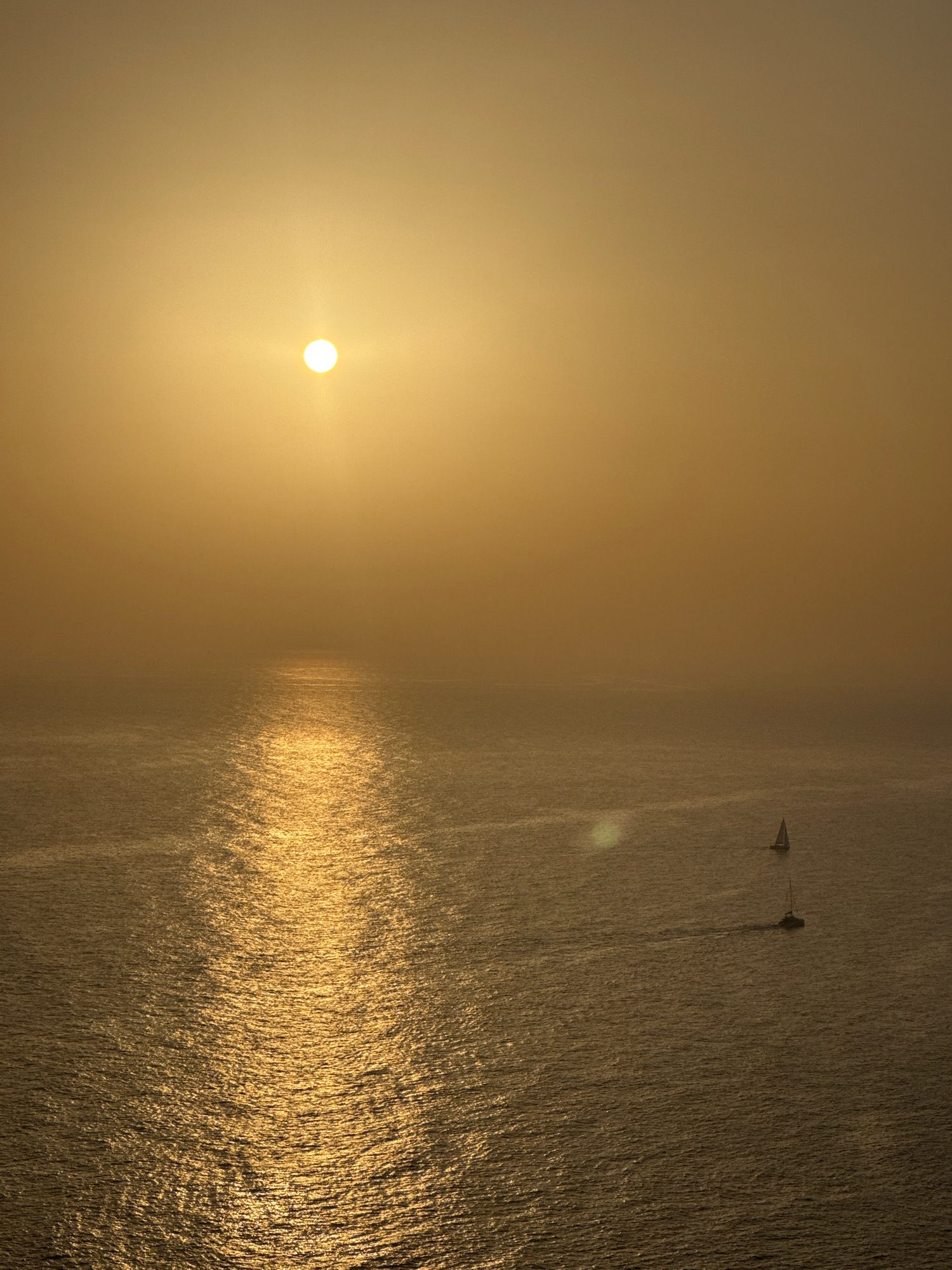 The image shows a wide shot of the Adriatic Sea at sunset, with the sun a bright disk low in the sky, casting a golden reflection across the water. Two sailboats are visible in the distance, one with sails up and the other with sails down. The sky is hazy and a warm golden-orange color. The foreground consists of choppy water with sunlight glinting off the surface. There is no indication of Zagora, Montenegro in the image.
