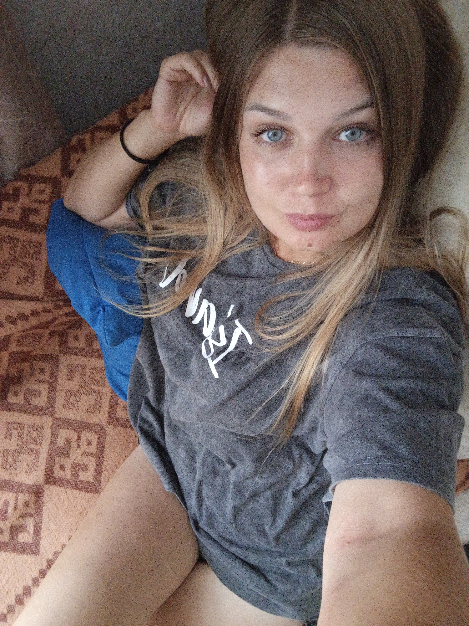 The image shows a close-up, overhead view of a young Caucasian woman lying on her back. She has fair skin, blue eyes, and long, blonde-brown hair that is partially covering her face and shoulders. She is wearing a grey t-shirt with white text on it, and a black wristband on her left wrist. Her right arm is bent at the elbow, with her hand resting on her head. She is lying on a bed with a patterned orange and brown bedspread. A blue pillow is visible to her left. The lighting suggests it is daytime, possibly indoors. The image is a selfie taken by the woman herself. There is no specific information within the image to indicate the location in Zlatoust, Russia.