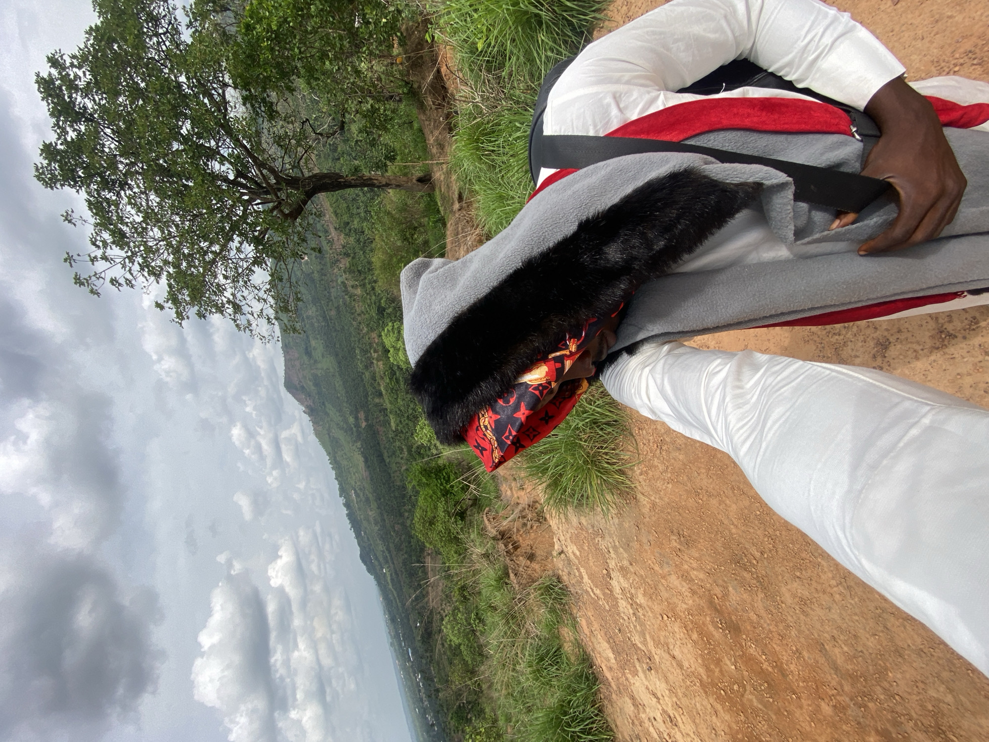 A person is reclining or resting on the reddish-brown earth of a hilltop, surrounded by patches of green grass and overlooking a vast natural landscape. The individual is dressed in a white long-sleeved top and white trousers. A gray blanket or garment with black faux fur trim covers part of their upper body. They wear a distinctive red patterned head covering, possibly a scarf or durag, featuring a pattern similar to the Louis Vuitton monogram. A dark-skinned hand rests on the gray garment.
In the midground, a prominent tree with lush green foliage stands, adding to the natural setting. The background reveals a panoramic view of distant hills or mountains blanketed in dense vegetation, stretching beneath an overcast but bright sky with scattered clouds. The scene suggests a daytime setting, possibly late morning or early afternoon. The overall atmosphere is one of peaceful relaxation, with the individual enjoying the expansive view from this high vantage point. The location is set in Amaigbo, Nigeria. No explicit readable text is visible in the image.