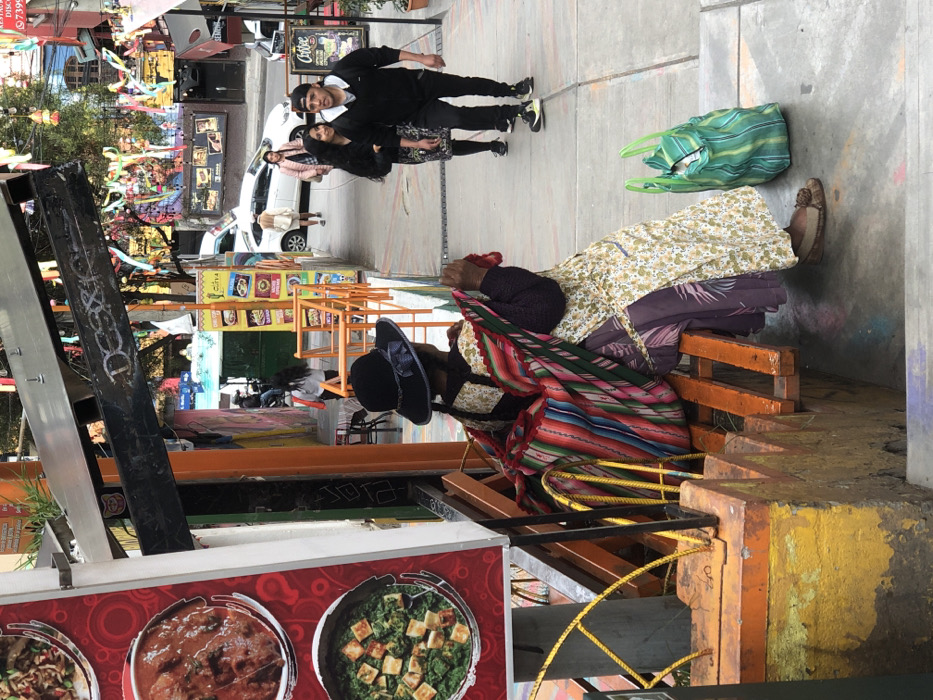 A street scene features a vendor seated on orange steps, adorned in a black hat and colorful textile, with a yellow floral dress visible. Overhead, a young couple in dark clothing walks past a green striped bag on the paved ground. In the background, pedestrians stroll by a white car and shops, decorated with colorful flags. A food stall's menu with images of Indian dishes is in the foreground.