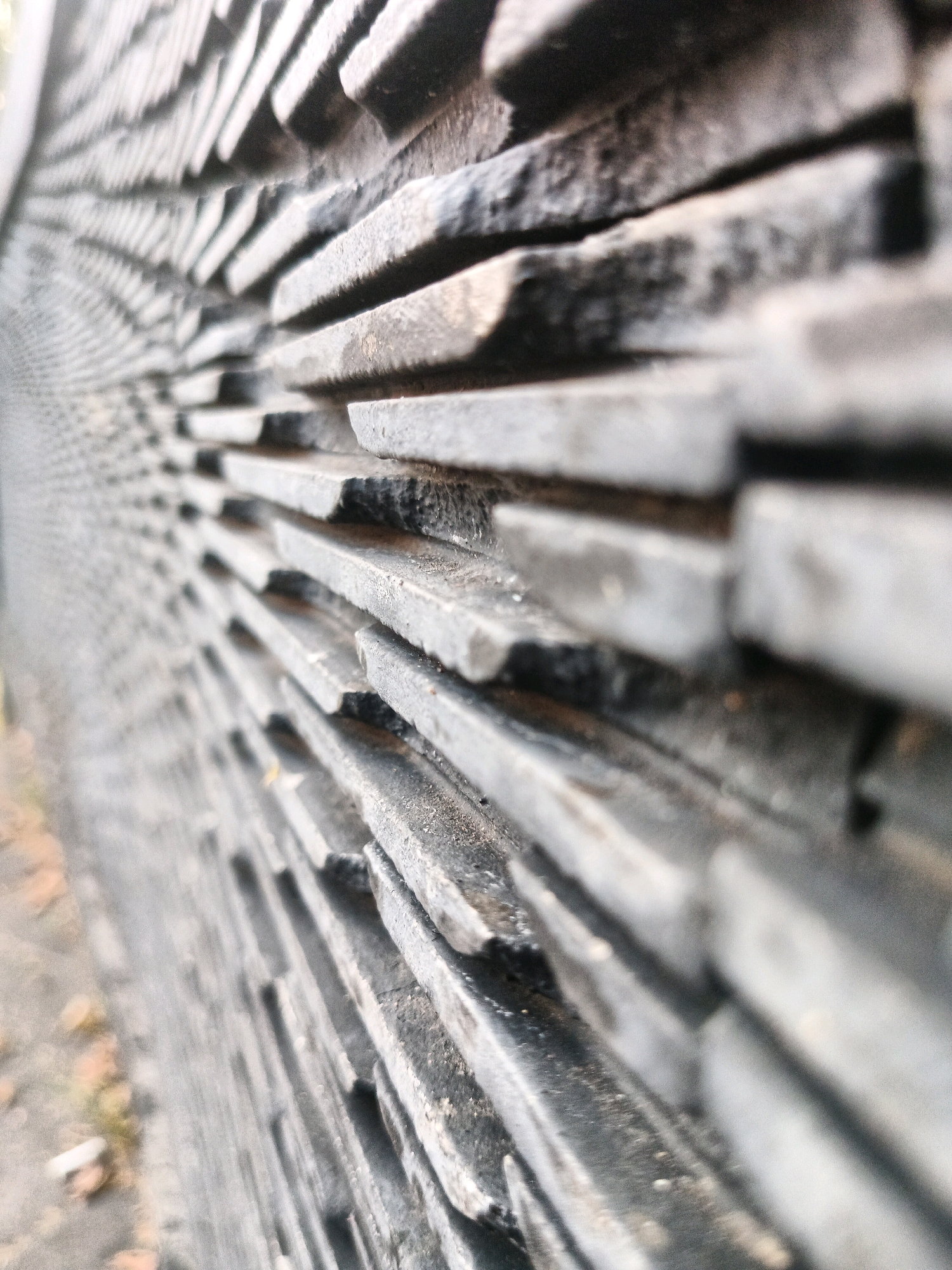 The image is a close-up, low-angle shot of a textured, dark gray stone wall, possibly slate, with a pattern of horizontal, overlapping rectangular stones. The stones have a rough, slightly weathered surface. The wall recedes into the background with a slight curve, creating a sense of depth. The lighting suggests an outdoor environment, with some areas of shadow and highlight on the stone surfaces. The focus is sharp on the stones in the foreground and middle ground, with the background becoming increasingly blurred. A small area of dry, fallen leaves and some debris is visible on the ground to the left of the wall, suggesting an exterior setting. No people or specific landmarks that would indicate a precise location in Bandung City, Indonesia are visible.
