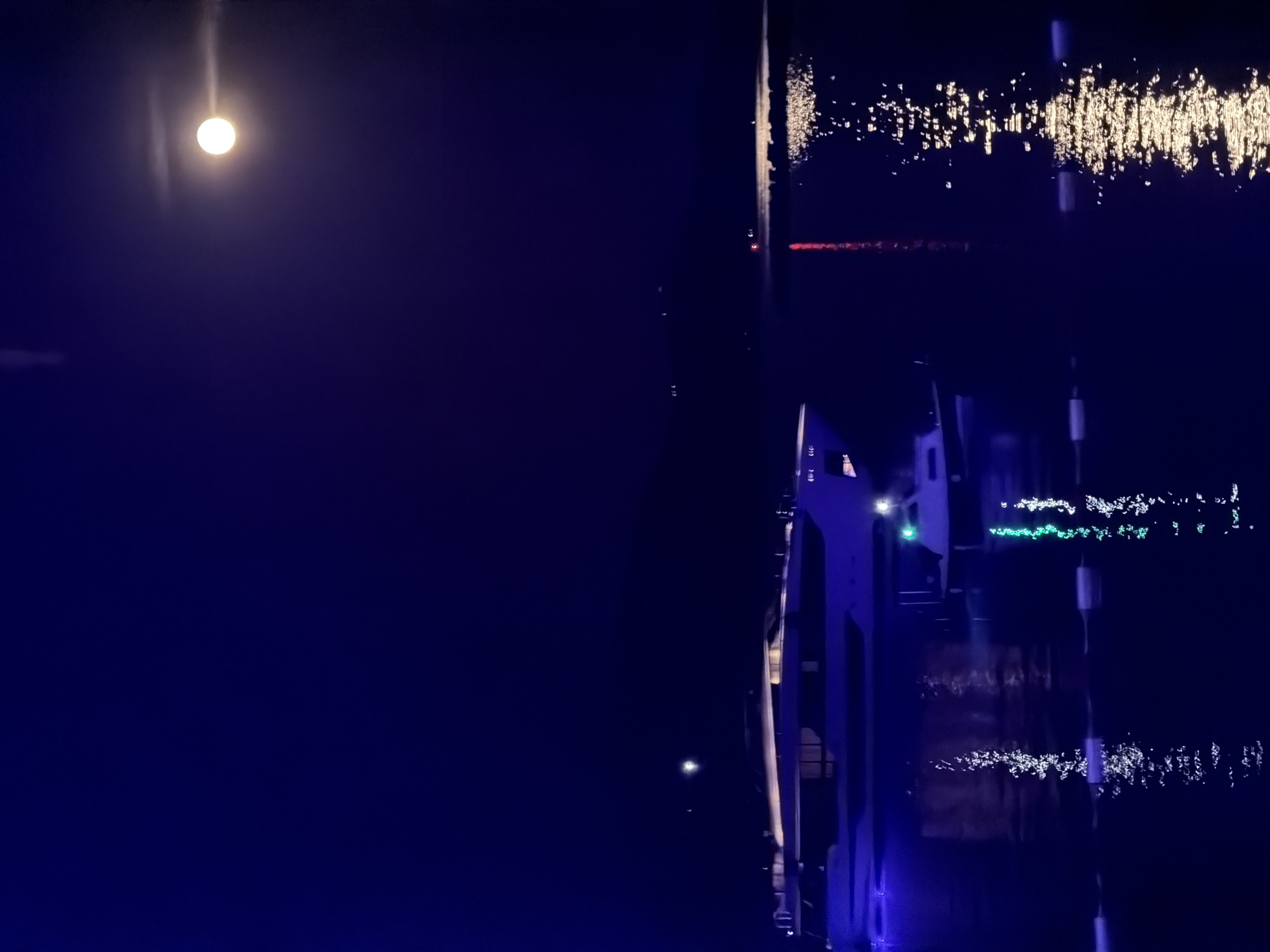 The image shows a night scene with a bright moon in the sky over Luštica, Montenegro. A large structure, possibly a bridge or a building with vertical supports, is visible on the right side of the frame, illuminated with blue and white lights. There are reflections of lights on the water's surface. In the distance, there are more lights, possibly from other buildings or boats.