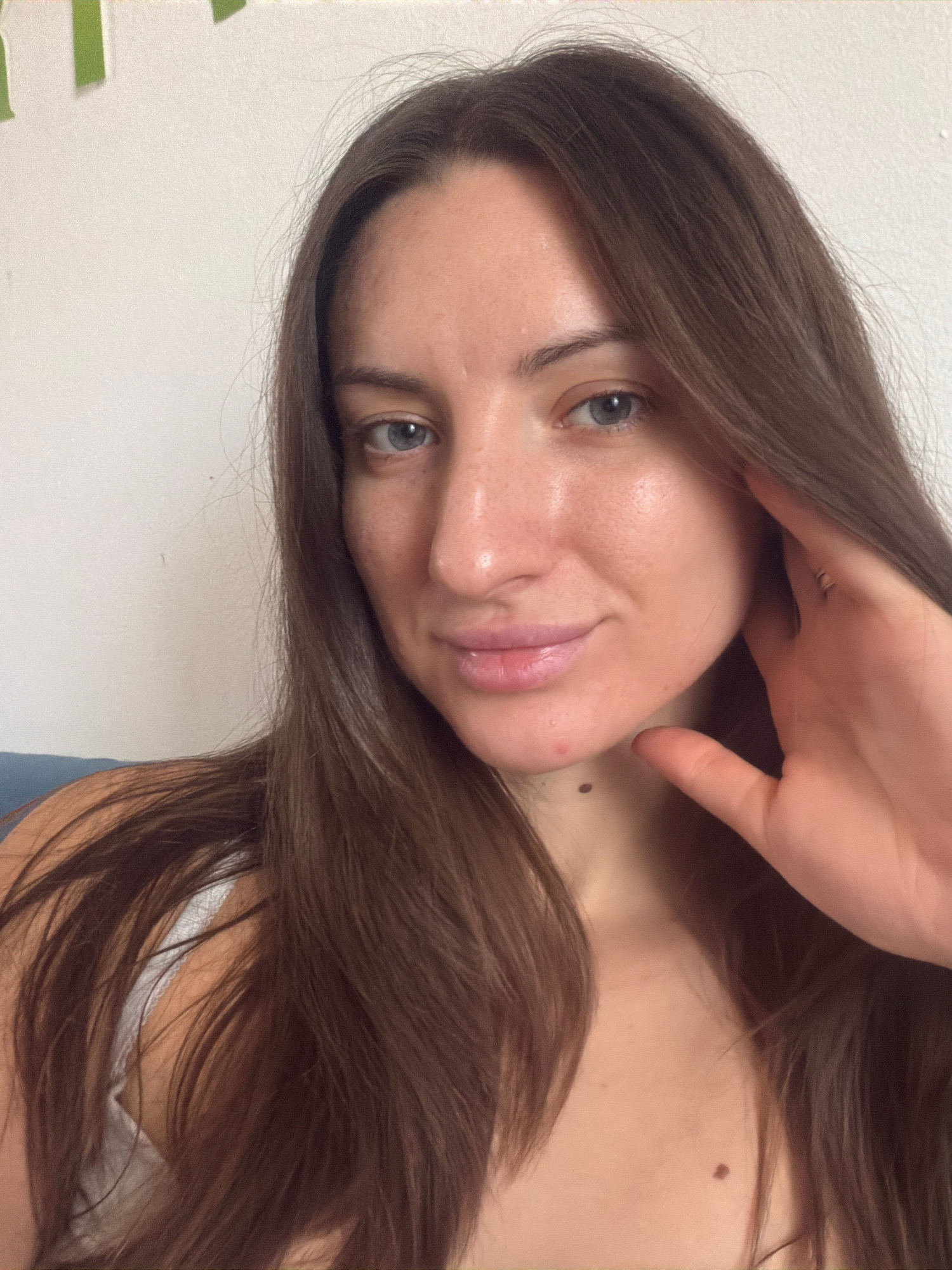 The image shows a close-up, waist-up, indoor portrait of a young woman with long, dark brown hair and blue eyes. She is looking towards the camera with a slight smile. Her left hand is raised to her face, touching her cheek. She is wearing a white top. The background is a plain white wall with some green decorative elements visible at the top left corner. The overall lighting is soft and natural. The image does not provide any information about the location being Sharm El Sheikh, Egypt.