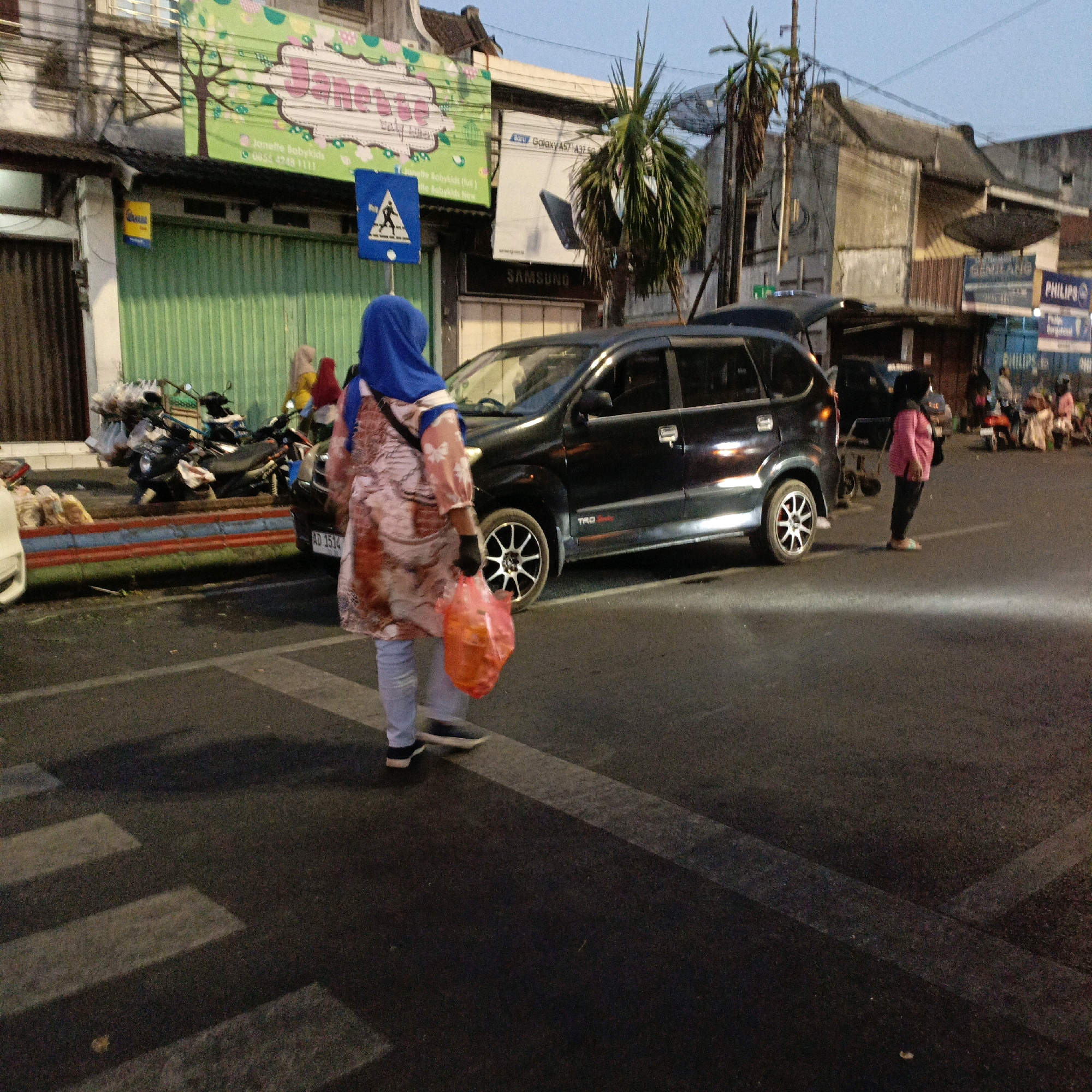 The image depicts a street scene in Salatiga, Indonesia, likely during the late afternoon or early evening, given the lighting conditions. In the foreground, a person wearing a blue hijab and a patterned long-sleeved top is walking across a pedestrian crossing, carrying a red plastic bag in their right hand. They are wearing light blue trousers and dark shoes. To their left, a black multi-purpose vehicle (MPV) is parked partially on the road. Behind the pedestrian crossing, several motorcycles are parked along the sidewalk, some with items piled on them.
The background shows a row of buildings, some of which appear to be shops or commercial establishments. Signs are visible on the buildings, including one with a green background and stylized text, possibly indicating a business name. Other signs show brand names like "Samsung" and "Philips". Palm trees are also visible, characteristic of the tropical climate. The road surface is asphalt, and there are clear lane markings. The overall impression is a busy urban street with typical street-level commerce and transportation.