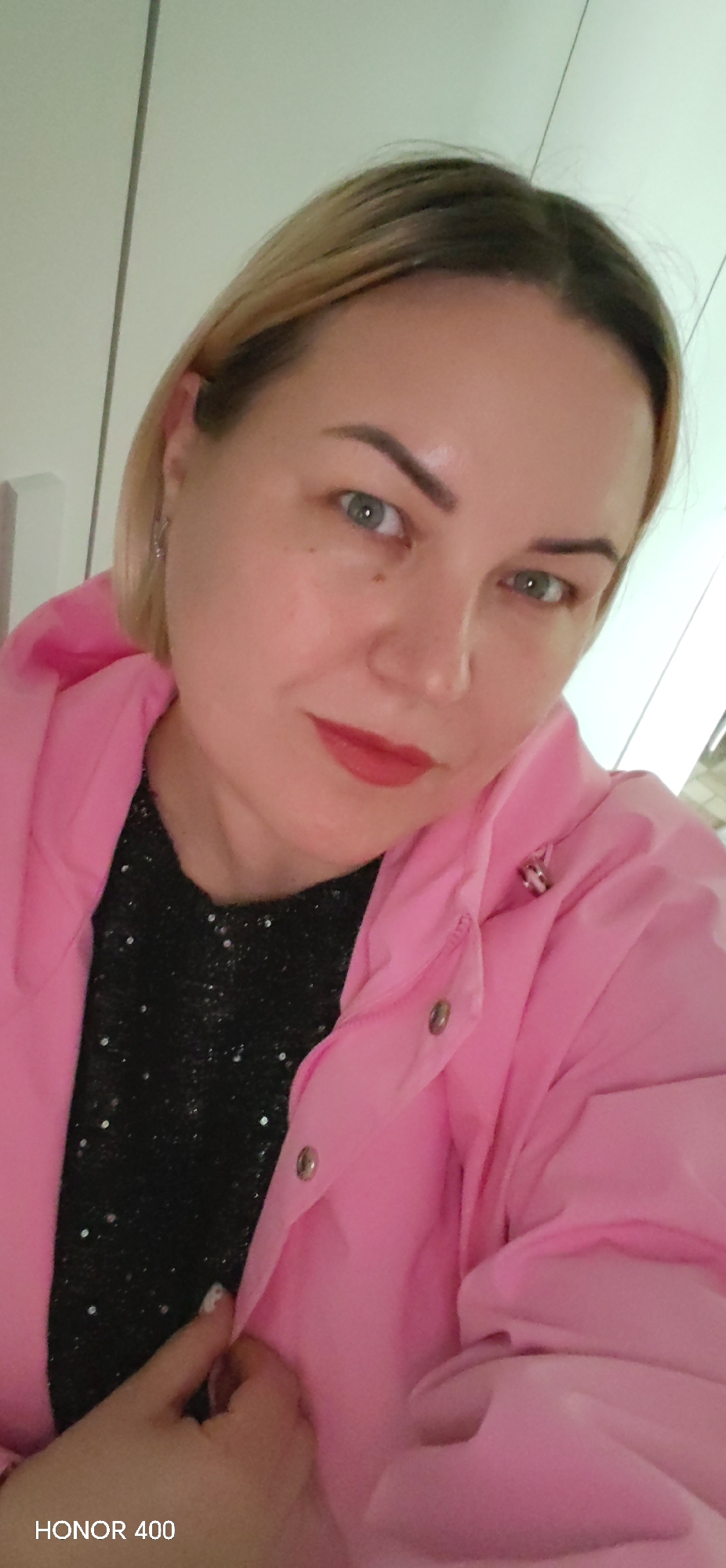 A close-up portrait captures a woman with light skin, short blonde hair showing darker roots, and green-blue eyes. She appears to be in her 40s. Her eyebrows are well-defined, and she wears red lipstick. In her visible left ear, there is a small silver earring. She is dressed in a bright pink hooded jacket, worn over a black top embellished with glitter or sequins. Her expression is calm and direct, with a subtle, gentle gaze towards the camera. The background consists of a plain, light-colored wall or cabinet, indicating an indoor setting. A silver snap button is visible on her pink jacket. In the bottom left corner of the image, the text "HONOR 400" is visible, likely a camera watermark. The image is taken in Noviy, Russia.