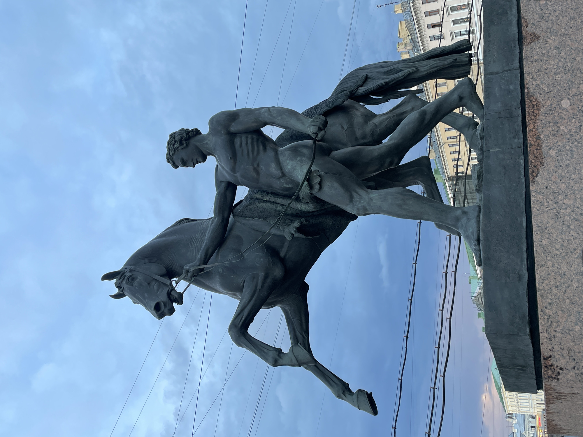 This is a photograph of the Anichkov Bridge in Saint Petersburg, Russia, featuring one of its famous equestrian sculptures. The sculpture depicts a muscular, nude male figure struggling to control a rearing horse. The horse is dark bronze, with its front legs raised dramatically in the air. The man is also bronze, with a lean physique, holding the horse's reins with his left hand and a whip or similar object in his right. His body is turned towards the horse, and his expression is one of effort and determination. The sculpture stands on a granite pedestal, which is partially visible at the bottom of the frame. In the background, the sky is overcast with a mix of blue and gray clouds. Several power lines stretch across the upper portion of the image. Behind the sculpture, the ornate facades of buildings in Saint Petersburg can be seen, characterized by their neoclassical architecture and mansard roofs. The overall lighting suggests it is either early morning or late evening.