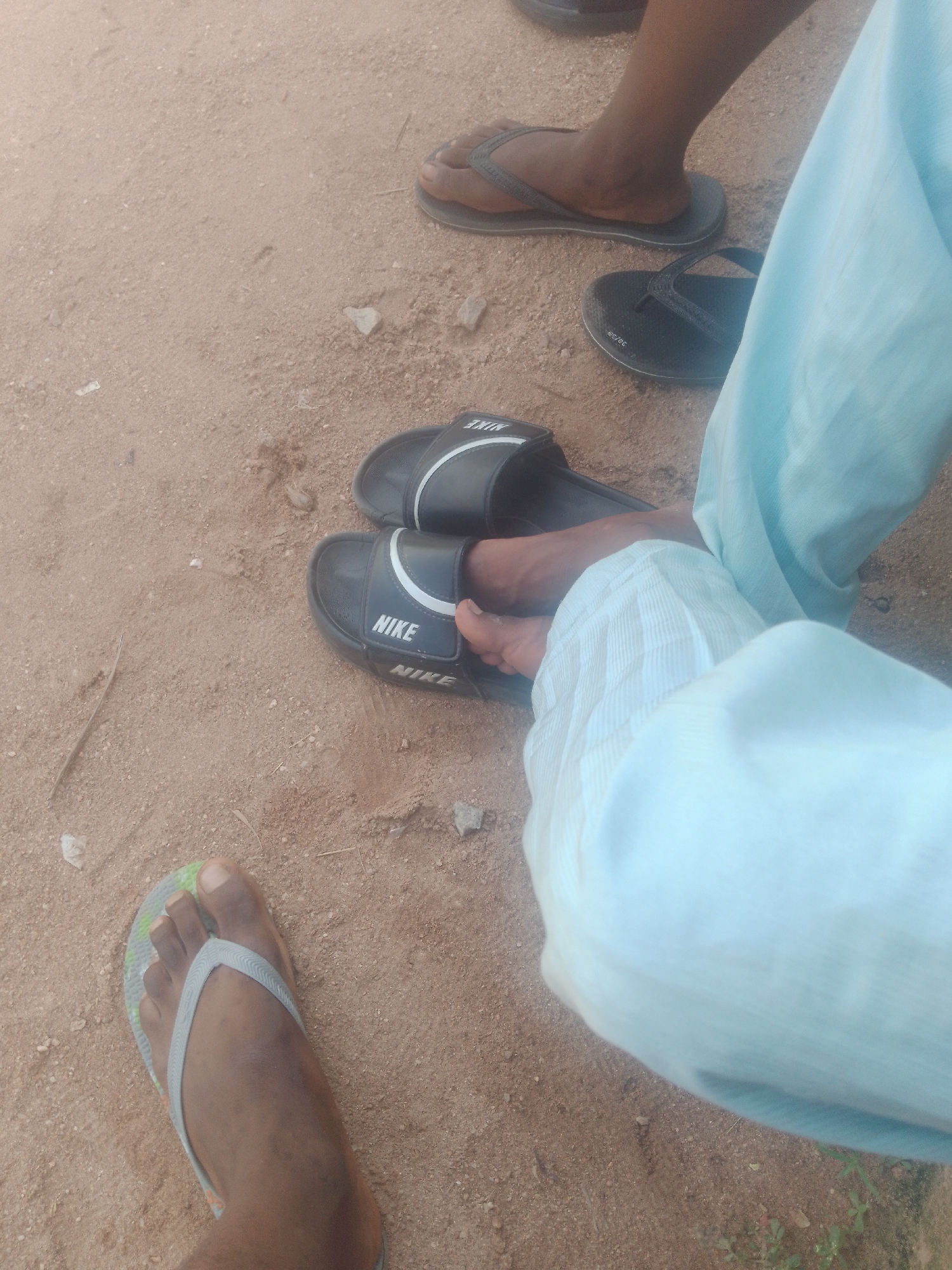 The image shows a close-up view of several pairs of footwear on sandy ground, likely in Jauro Yinu, Nigeria. In the foreground, a left foot wearing a gray and green flip-flop is visible. Behind it, a pair of black Nike slides is on the ground. Further back, another pair of black slides is also visible. The lighting suggests it is daytime, and the environment appears to be outdoors. The composition focuses on the footwear and the ground, with portions of light blue fabric from clothing visible on the right side of the frame.