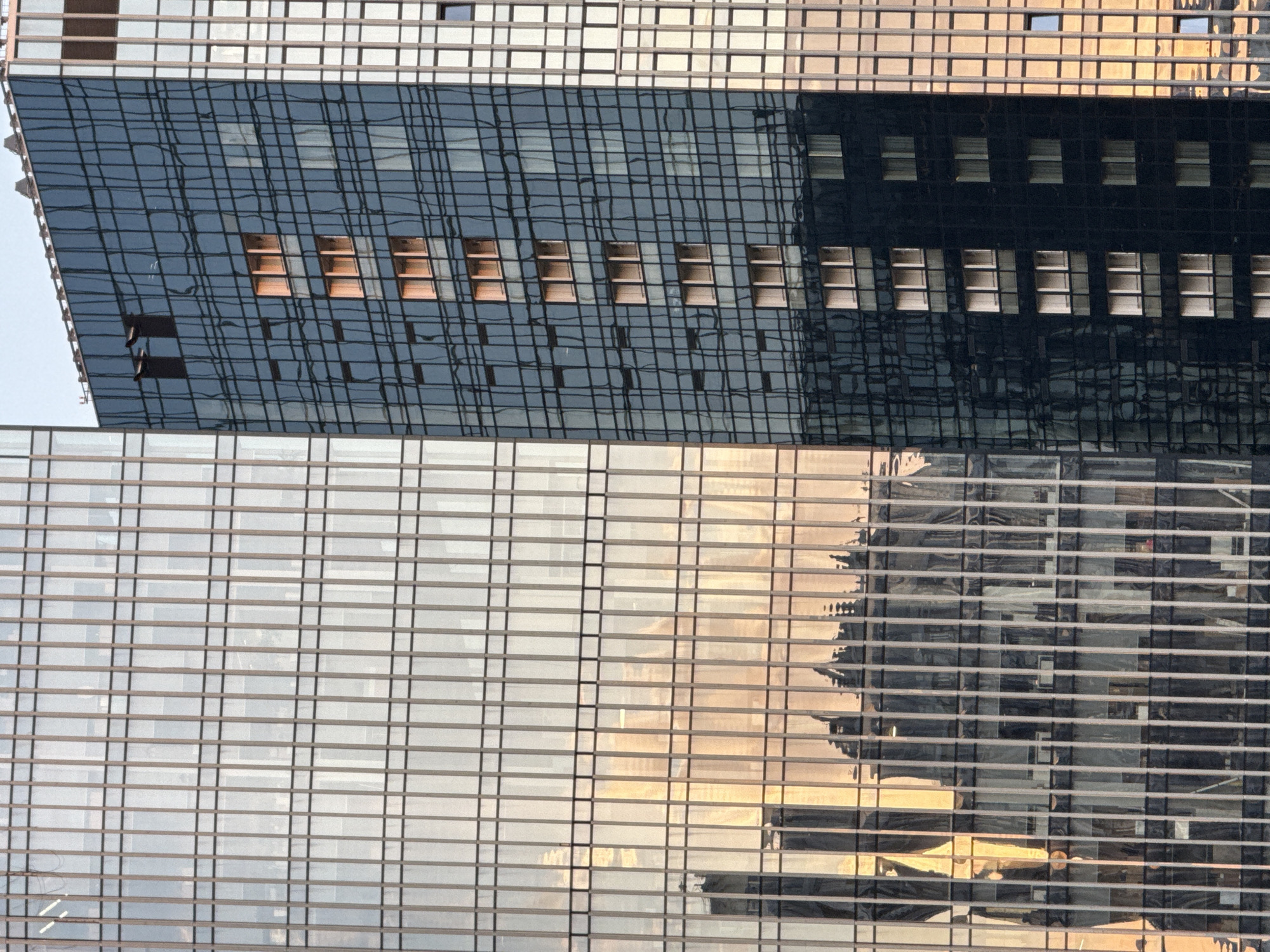 This media file is a vertical, close-up shot of modern architecture, specifically the exterior of a glass-faced skyscraper. The primary subjects are the building's facade, characterized by a grid-like pattern of windows and structural elements. The upper portion of the image reveals a dark, reflective glass surface, broken by a series of rectangular window openings that appear to be empty or dimly lit from within. Below this, a lighter, more translucent glass section dominates the lower half of the frame. This area reflects the warm, golden hues of what appears to be a sunset or sunrise, interspersed with darker shapes that are reflections of other buildings or structures.
The scene is abstract, focusing on the geometric patterns and reflections created by the building's design and its interaction with light. There are no people or discernible objects within the frame, other than the architectural elements themselves. The activity depicted is passive, simply the presence of the building and the interplay of light and reflection.
Notable details include the sharp, defined lines of the building's structure and windows, contrasted with the distorted, wavy reflections on the dark glass. The warm, golden light suggests a time of day at either dawn or dusk. The location context provided, Khimki, Russia, is not visually apparent from the image itself, as it offers no specific landmarks or cultural cues. There is no visible text in the media file. The overall impression is one of urban modernity and the aesthetic qualities of glass architecture.