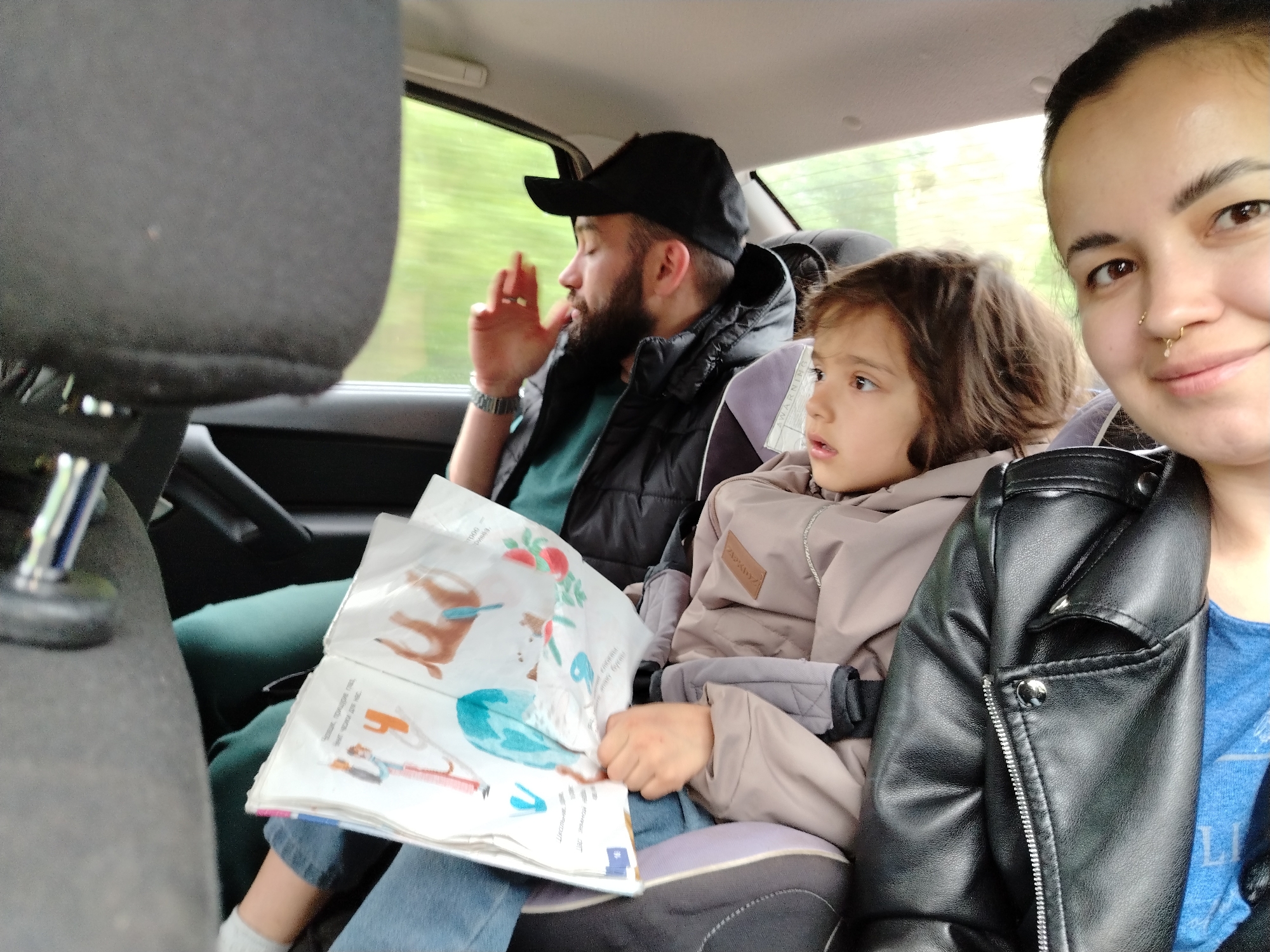A daytime, medium shot depicts three individuals inside a car, possibly traveling on a road. The scene appears to be set in Краснояр, Russia based on information provided. The individual on the right, closest to the camera, is a woman with dark hair pulled back, wearing a black leather jacket over a blue shirt. She is looking at the camera and smiling slightly. A child, possibly a girl, sits in the middle, looking to the left. The child is wearing a tan jacket and is partially buckled into a car seat. The person on the left, further back, is a man with a beard and a black cap. He is wearing a black vest over a dark green shirt and is looking out the window. The man is holding a book that is open, displaying colorful images, perhaps for the child. The interior of the car is visible, including the headrests and windows through which green foliage can be seen, indicating they may be traveling past trees or a forested area. The lighting suggests it is daylight but possibly overcast.