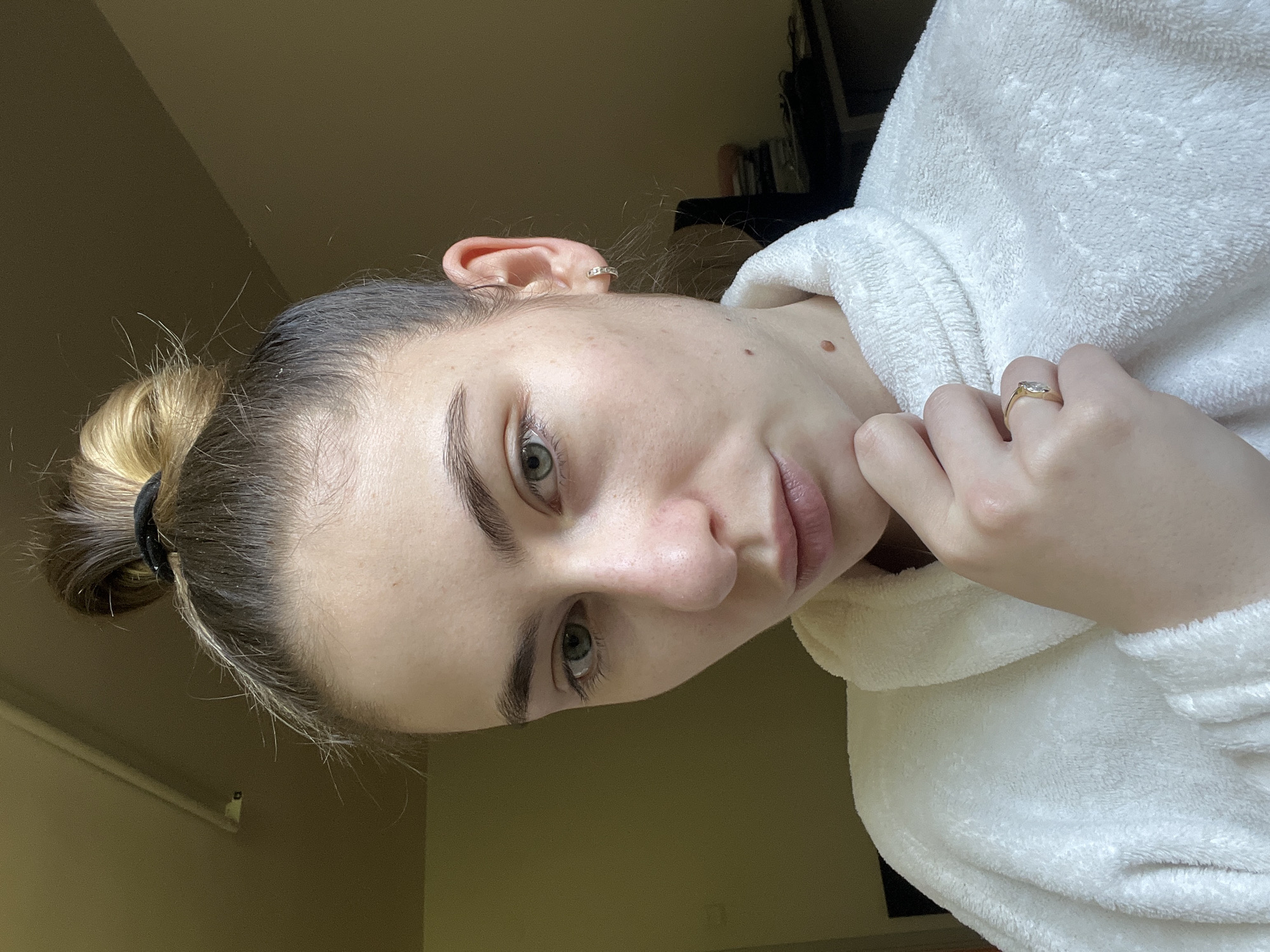 A young woman with light skin and green eyes, wearing a white fluffy robe and a gold ring on her left ring finger, is looking at the camera. Her hair is pulled back in a bun secured with a black elastic band. She has a small earring in her left ear. The background is a blurred room with a dark shelf and light-colored walls. The location is Unknown, Unknown.