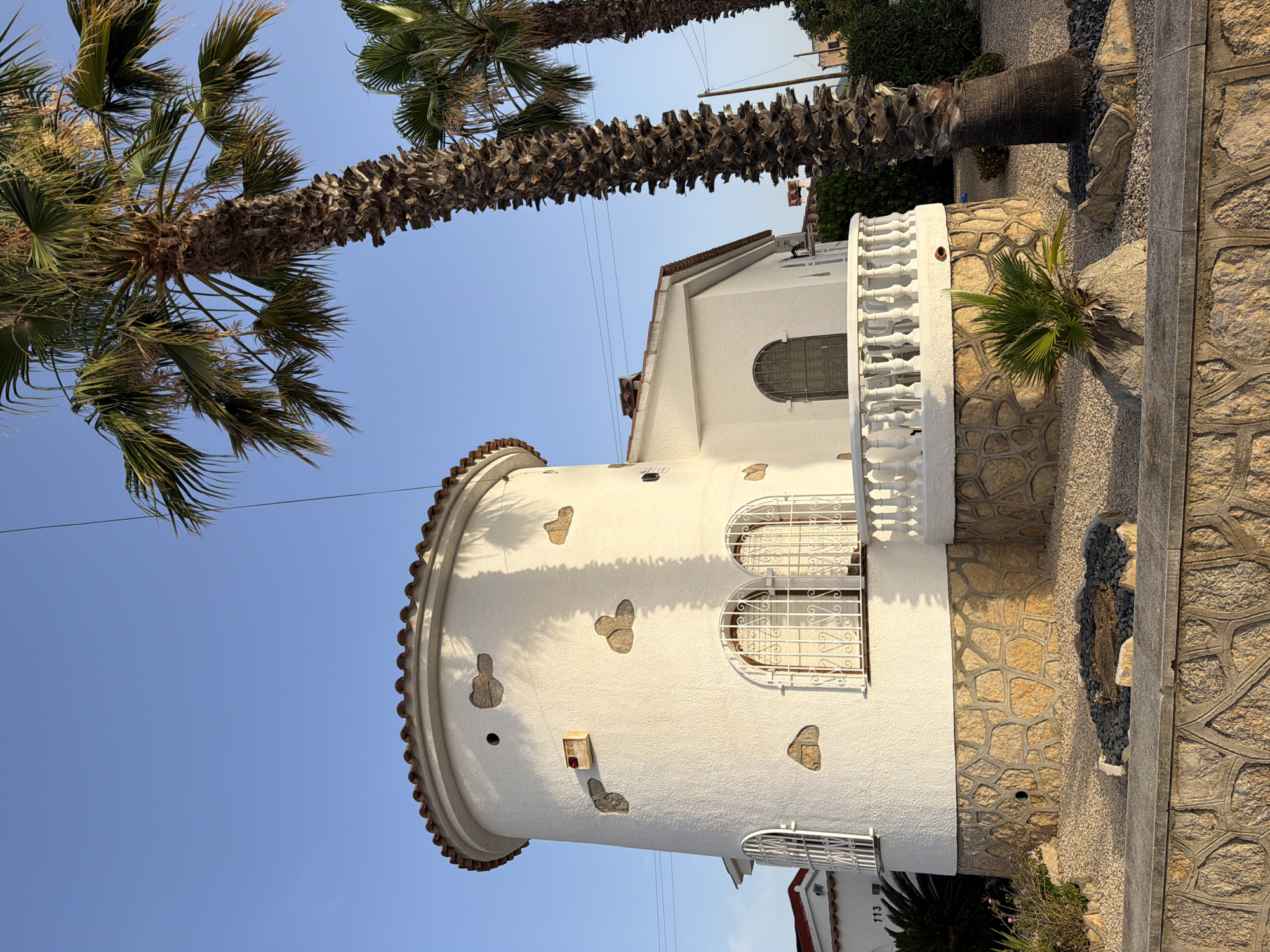 The image shows a white, cylindrical house with a red tile roof, reminiscent of a tower, located in Mont-roig del Camp, Spain. The lower portion of the house is clad in rough-hewn, tan-colored stones. Two arched windows with white decorative bars are visible on the cylindrical section. A white balustrade curves around a balcony extending from the house. The house is surrounded by gravel, with a small fire pit filled with dark stones and logs in the foreground. Several palm trees, including a tall one prominently positioned to the right of the house, are present. The sky is clear and blue. The overall scene depicts a residential building in a sunny, likely Mediterranean, environment.